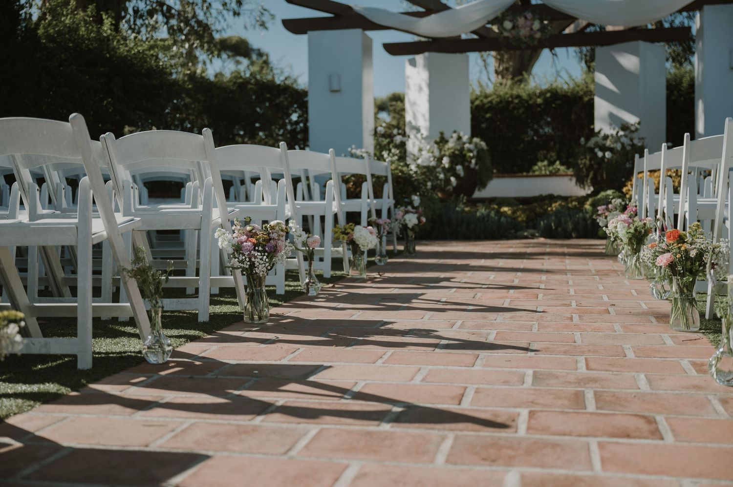 Outdoor wedding ceremony aisle with white folding chairs and colorful floral arrangements on brick pathway.