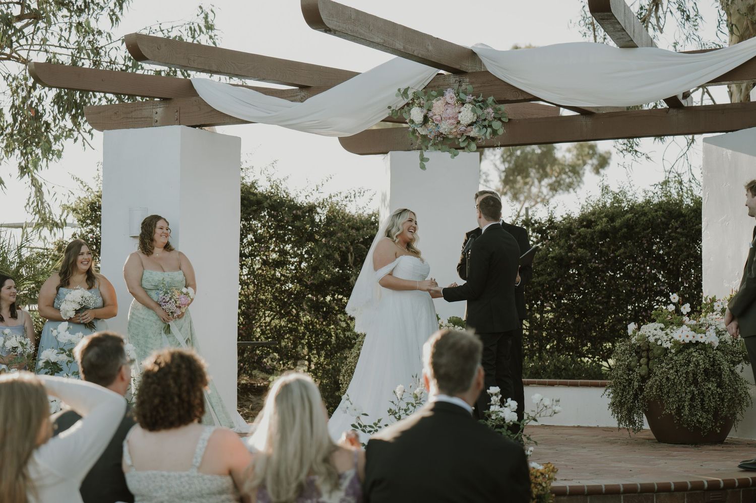 Bride and groom exchange vows at outdoor wedding ceremony with floral pergola and bridesmaids in sage green dresses.