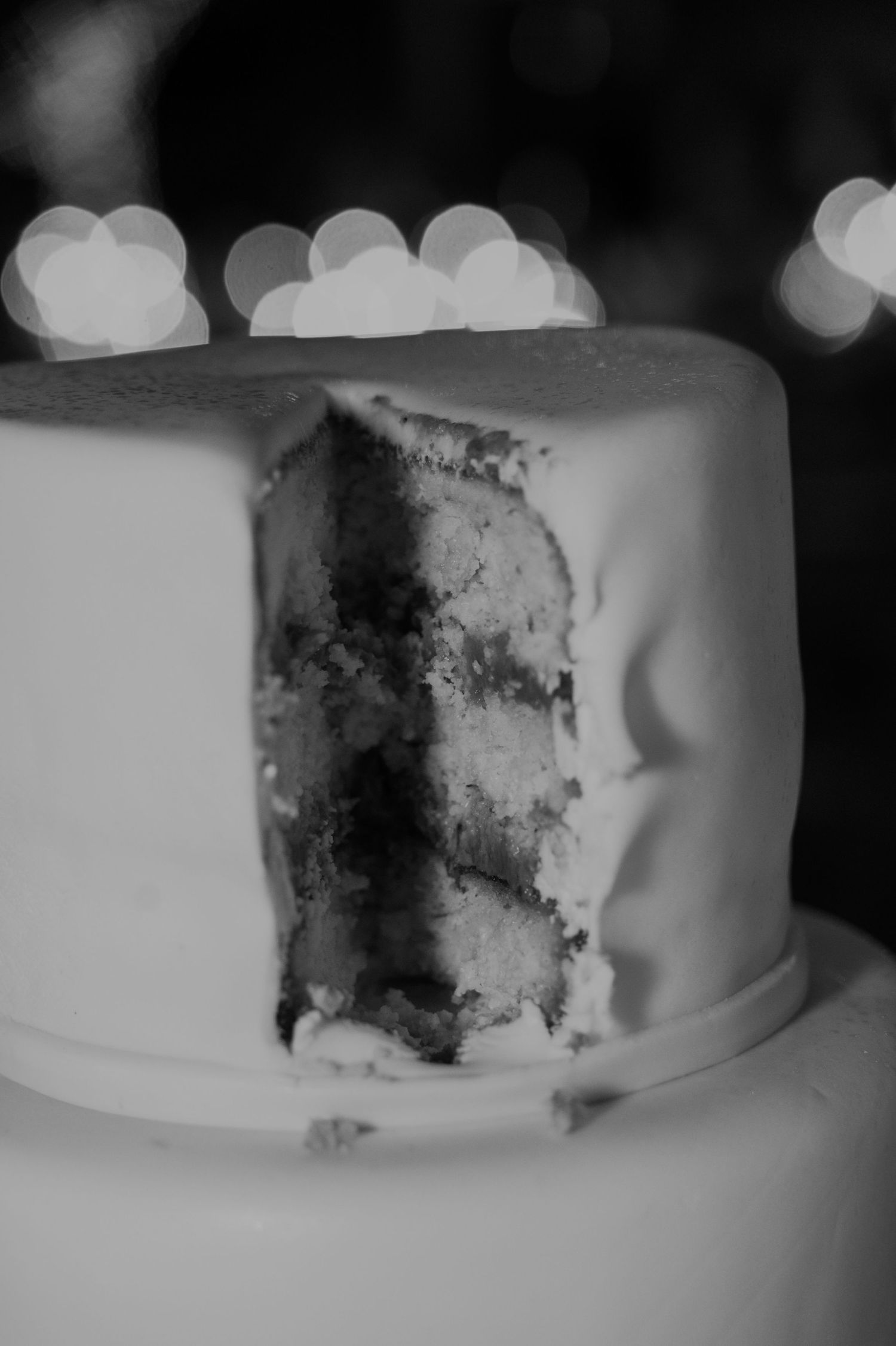 Black and white close-up of a sliced multi-layer wedding cake with bokeh lights in the background.