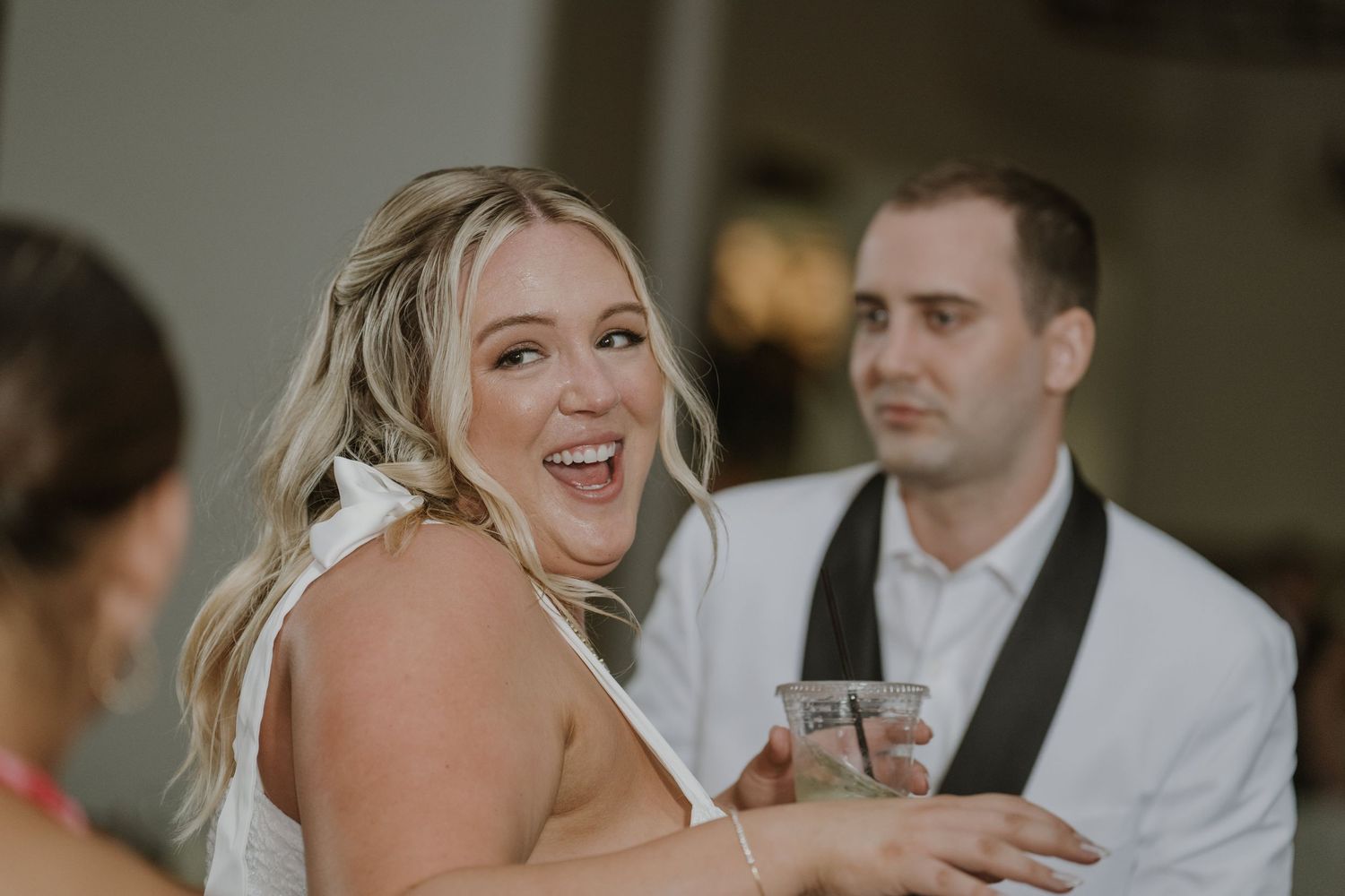 Joyful bride laughing and holding a drink at her wedding reception with groom in background.