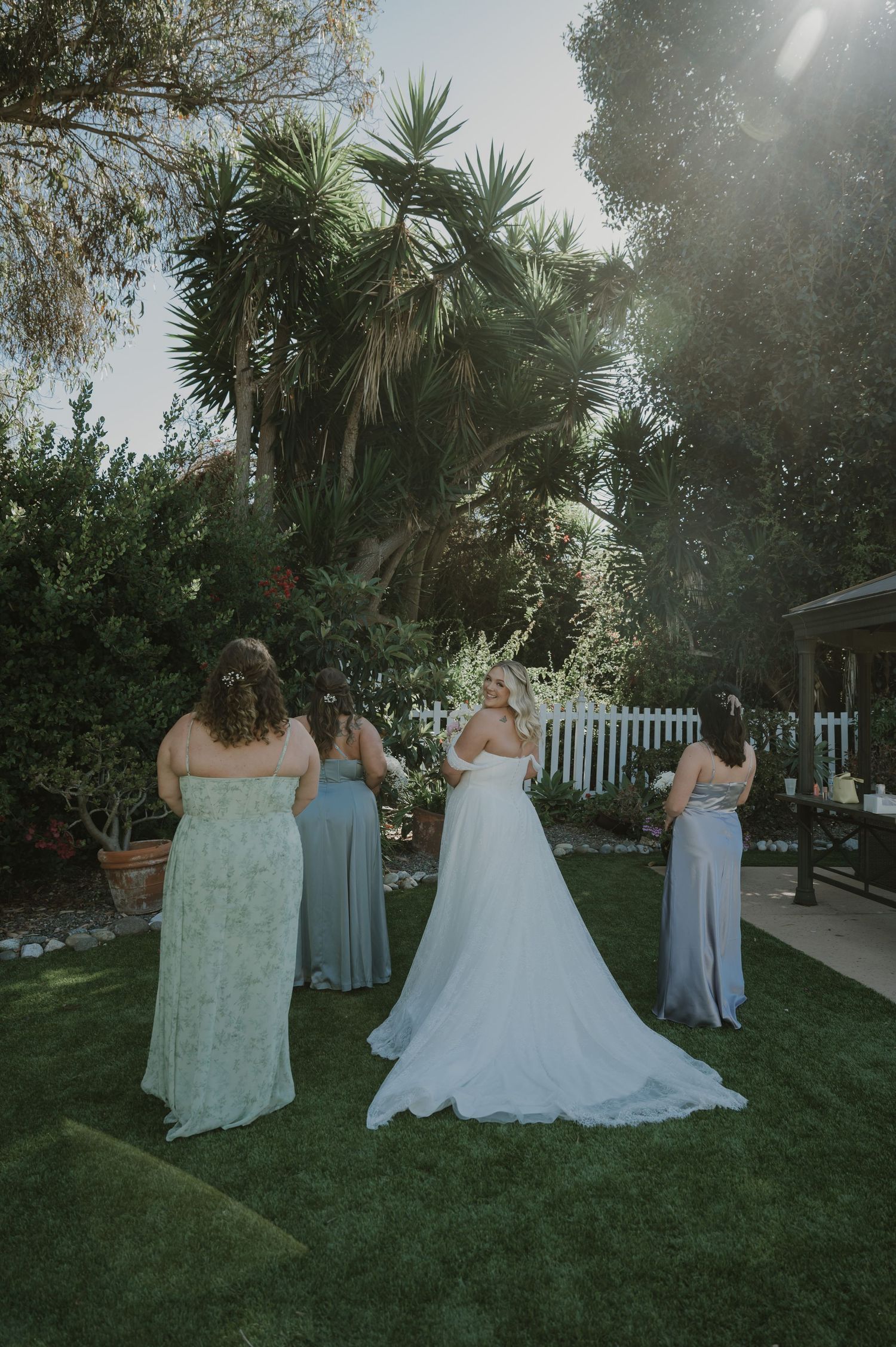 Bride in white gown stands with bridesmaids in sage dresses in sunny garden setting.