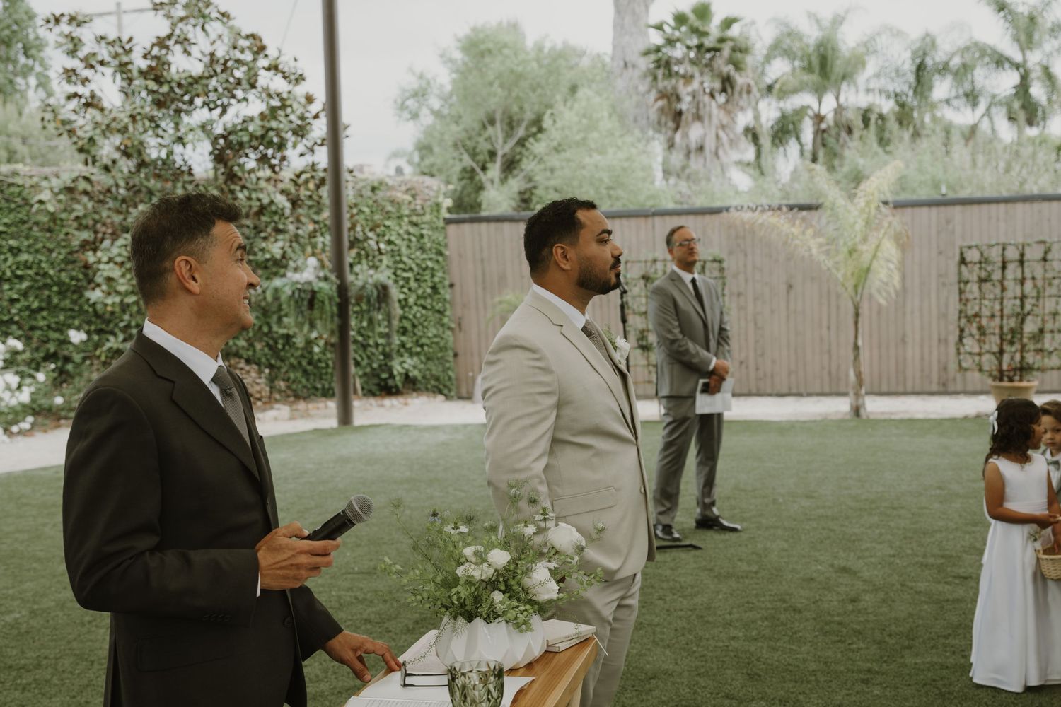 Groom in light gray suit stands at outdoor wedding ceremony on green lawn with floral arrangements.
