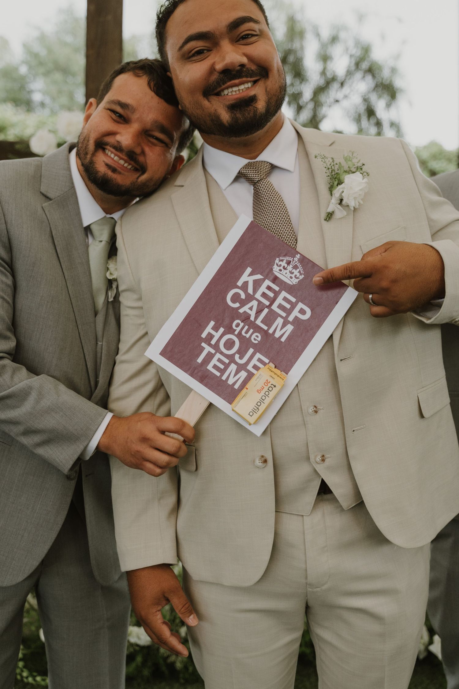 Two groomsmen in beige suits smiling and holding a Keep Calm wedding sign.