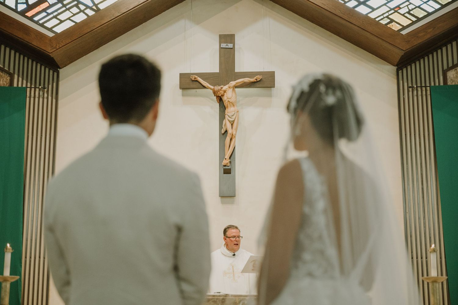 Bride and groom stand before officiant at church altar with crucifix, during their wedding ceremony.