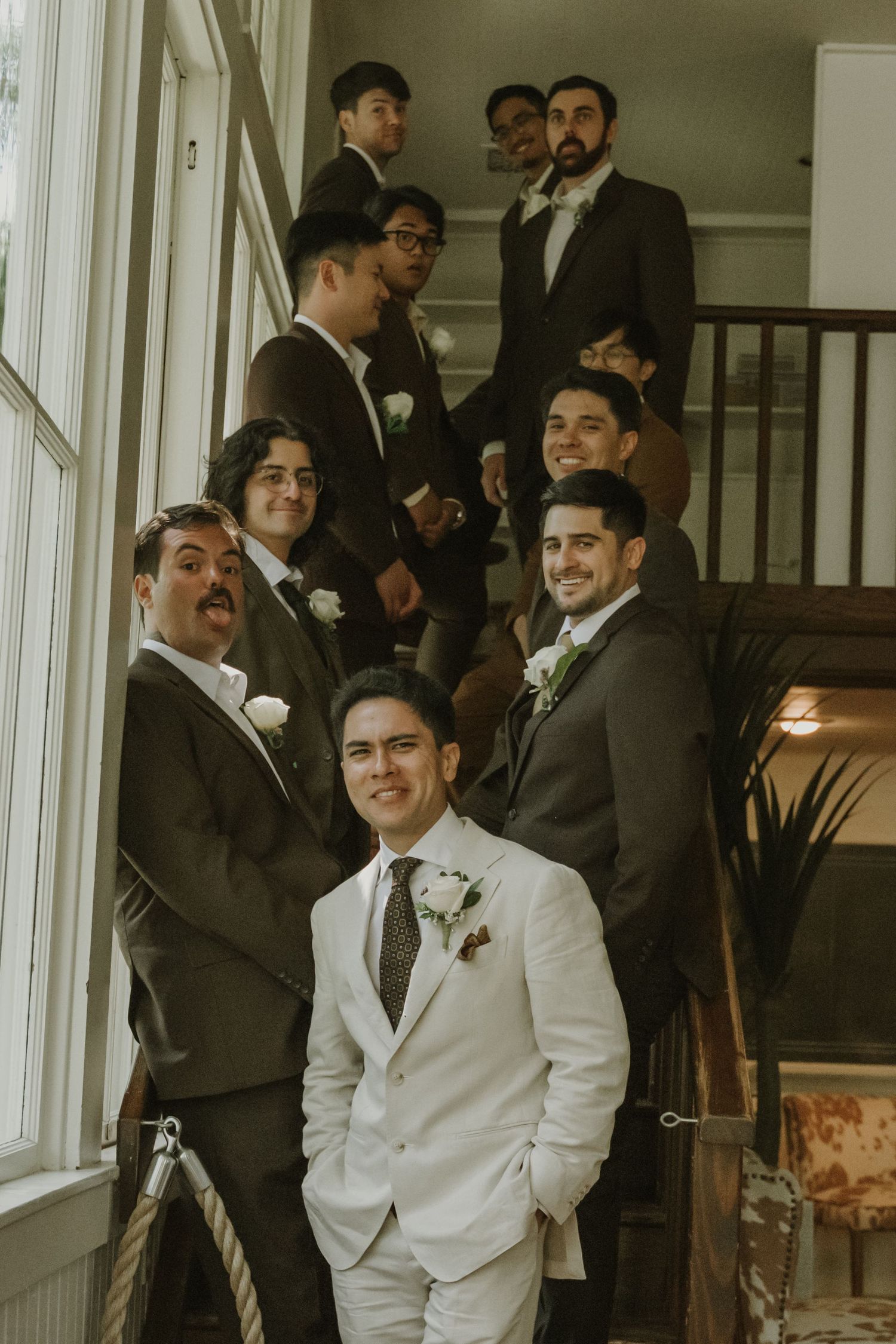 Smiling groom in ivory suit stands with groomsmen arranged on vintage interior staircase.