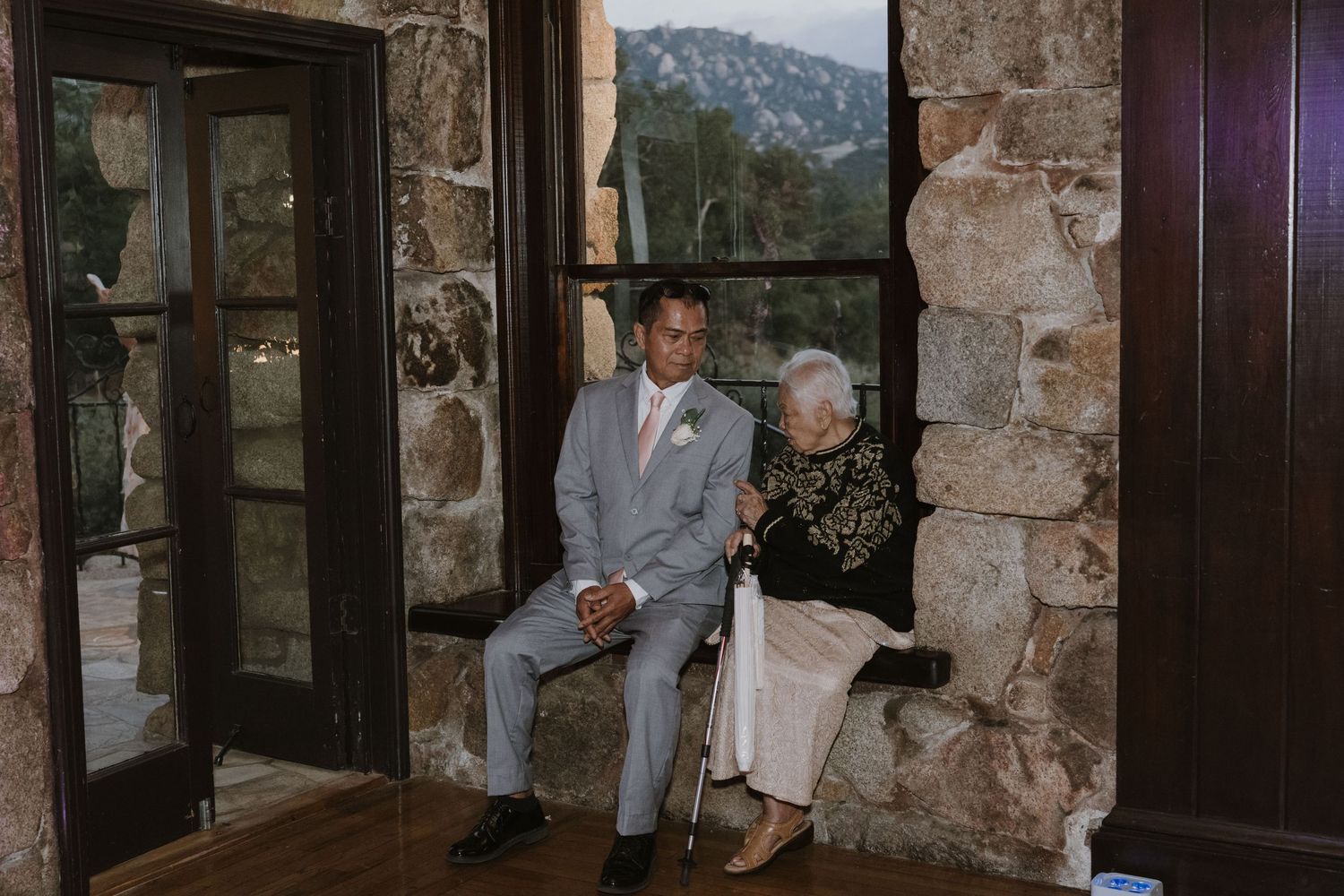 Groom in gray suit sitting with elderly grandmother at a rustic stone wedding venue.