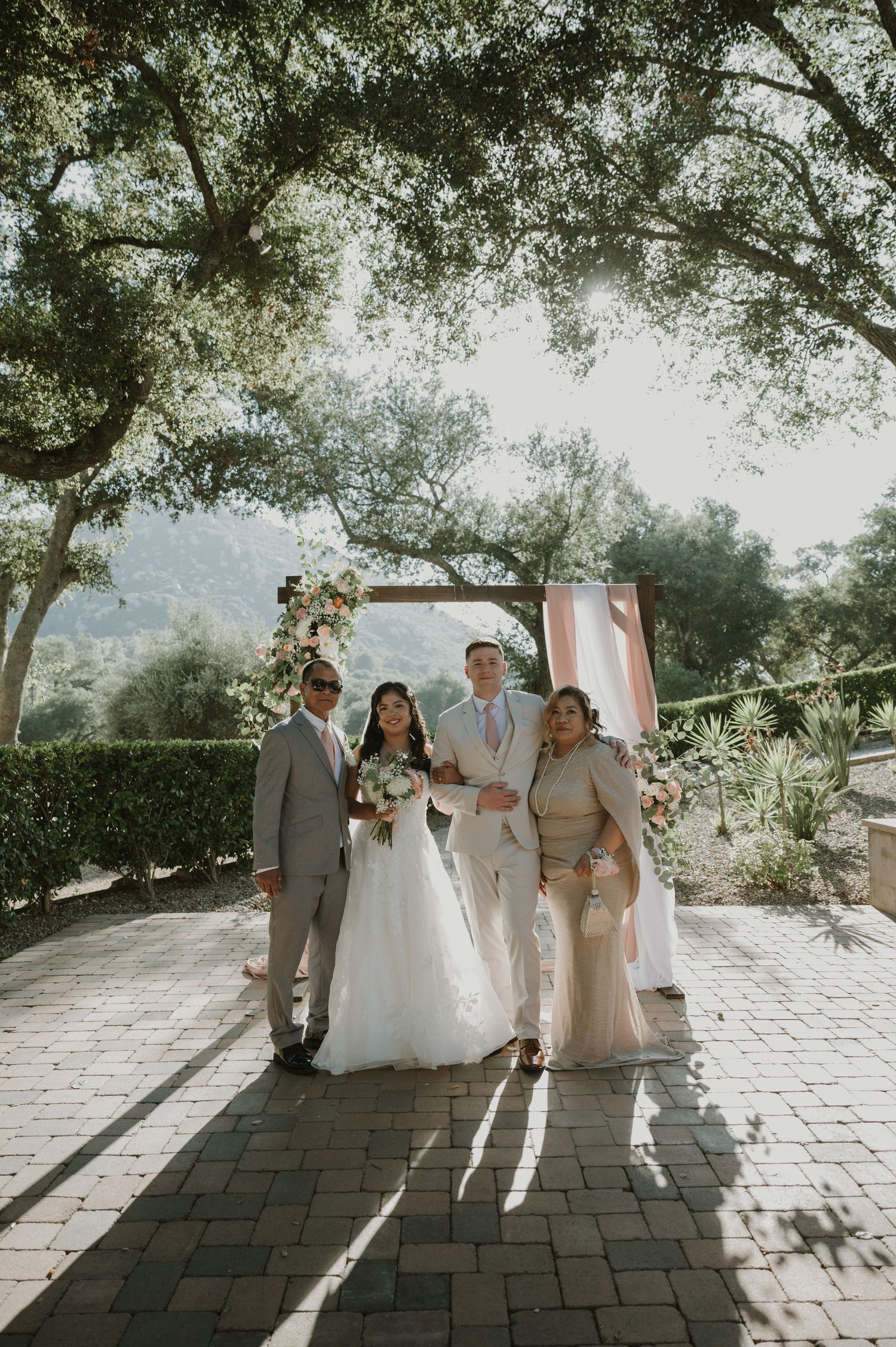 Newlyweds pose with family under tree canopy at outdoor wedding venue with mountain views.