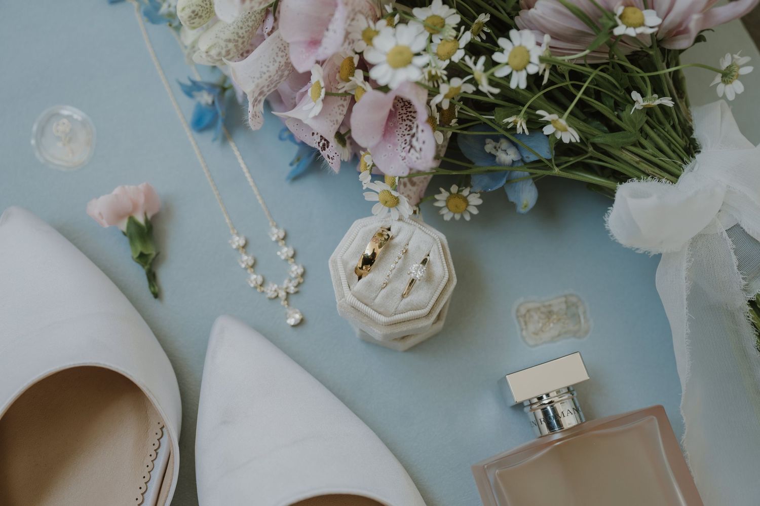 Elegant wedding flat lay with bridal heels, floral bouquet, ring box, pearl necklace, and perfume on blue background.