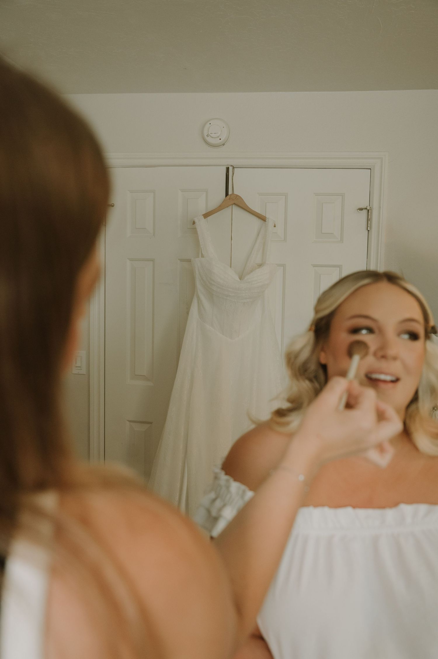 Blonde bride applying makeup in mirror with white wedding dress hanging in background.