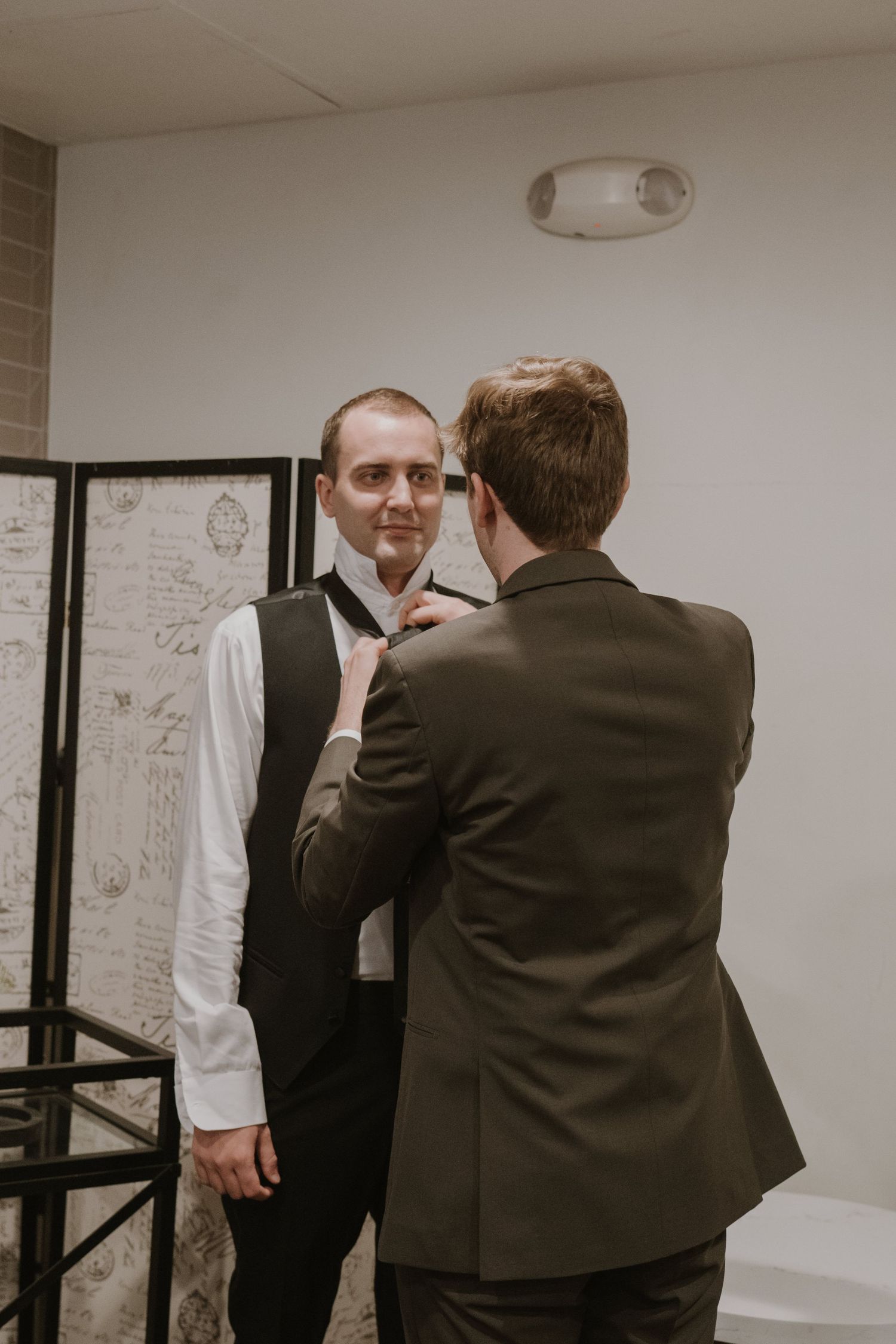 A groomsman adjusts another mans tie while getting ready for a wedding ceremony indoors.