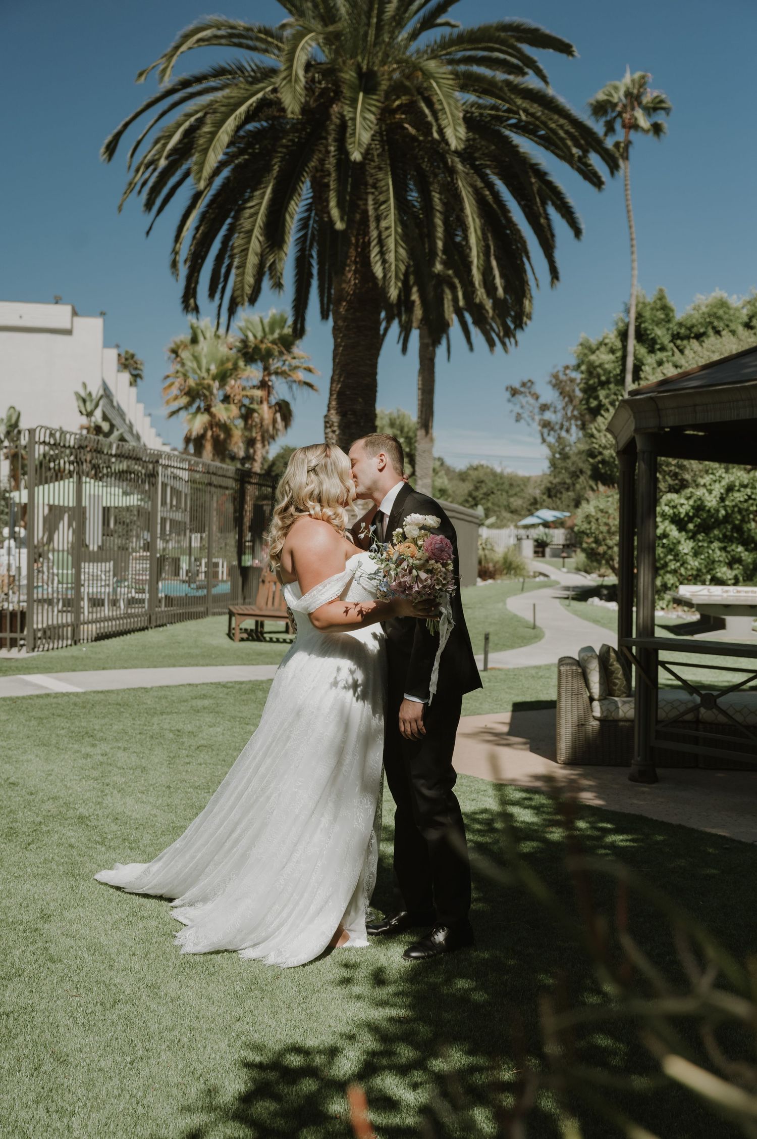 Bride and groom share a first kiss on a sunny lawn with tall palm trees at an outdoor wedding venue.