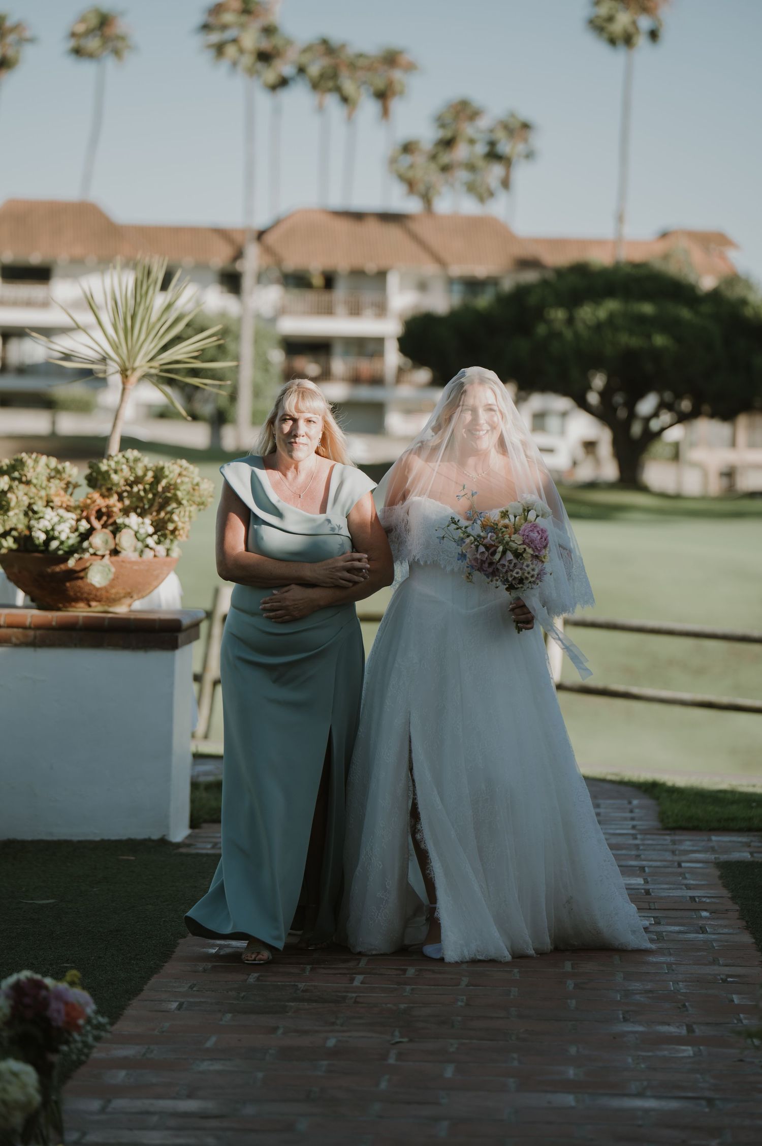 Bride in white gown and veil walks with woman in sage green dress past palm trees at outdoor wedding.