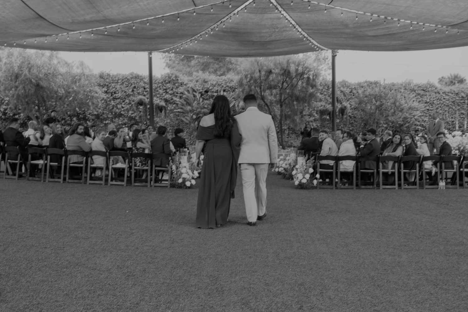 Black and white photo of couple walking toward outdoor wedding reception under tent with seated guests.