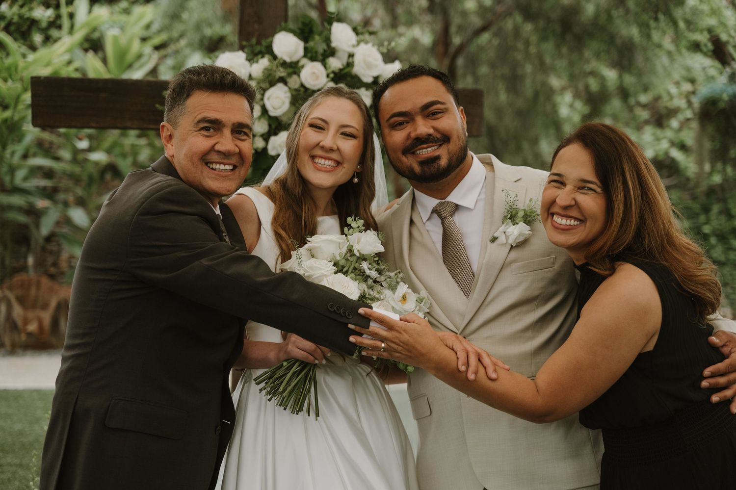 A joyful wedding party with bride in white gown holding bouquet, groom in tan suit, and two guests smiling outdoors.