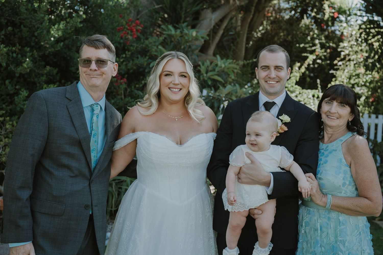 Bride and groom pose with family including a baby at an outdoor wedding ceremony surrounded by lush greenery.