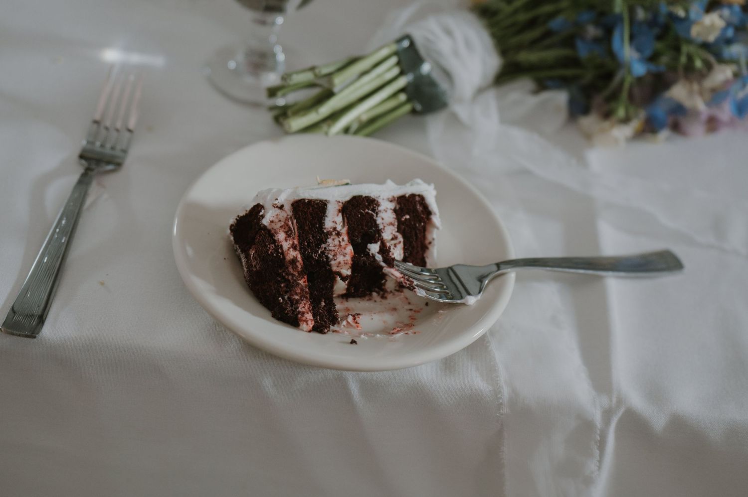 A slice of chocolate layer cake with white frosting on a white plate at a wedding reception table setting.