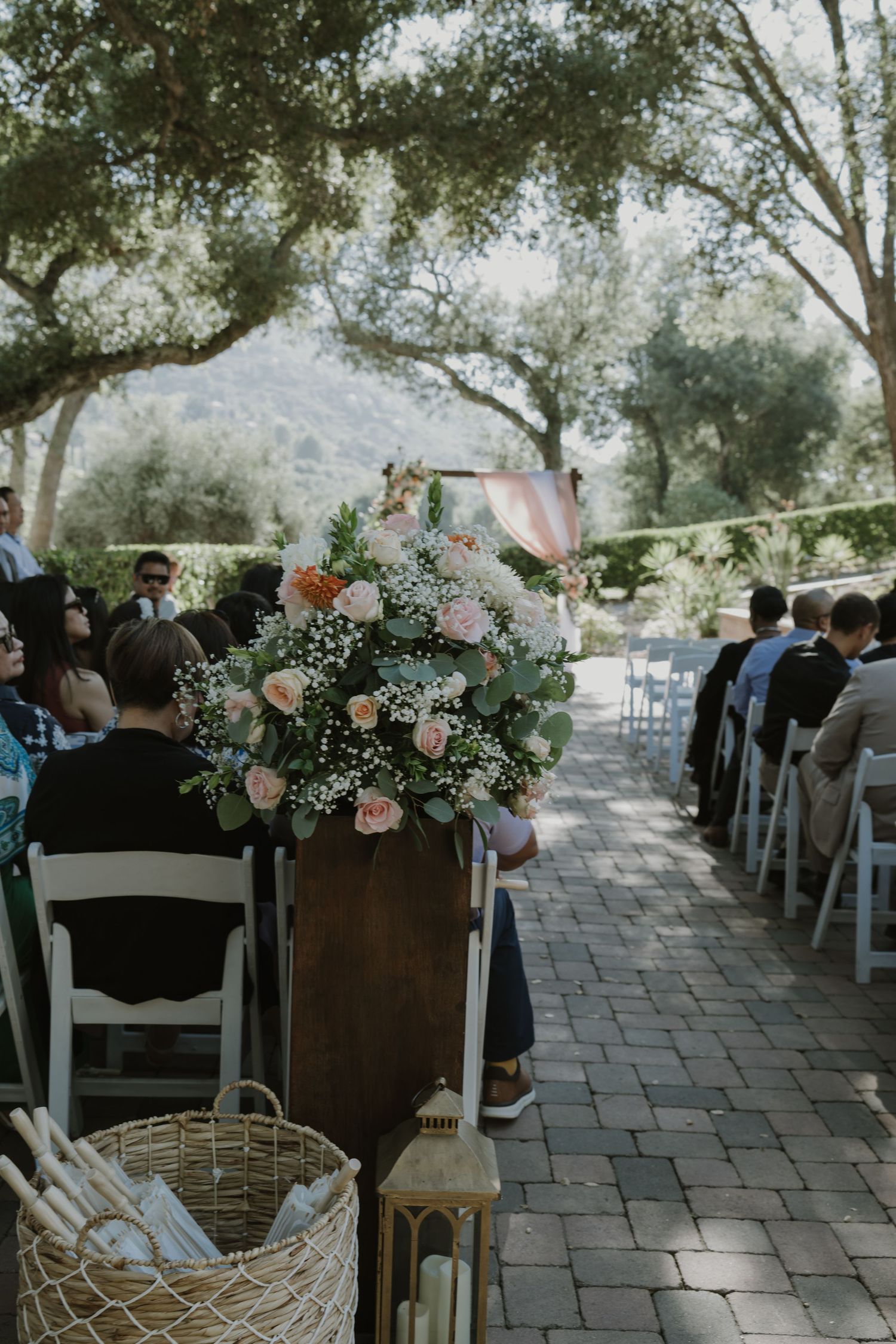 Outdoor wedding ceremony aisle with floral arrangements of pink roses and greenery on wooden pedestals, white chairs, and tree-lined path.