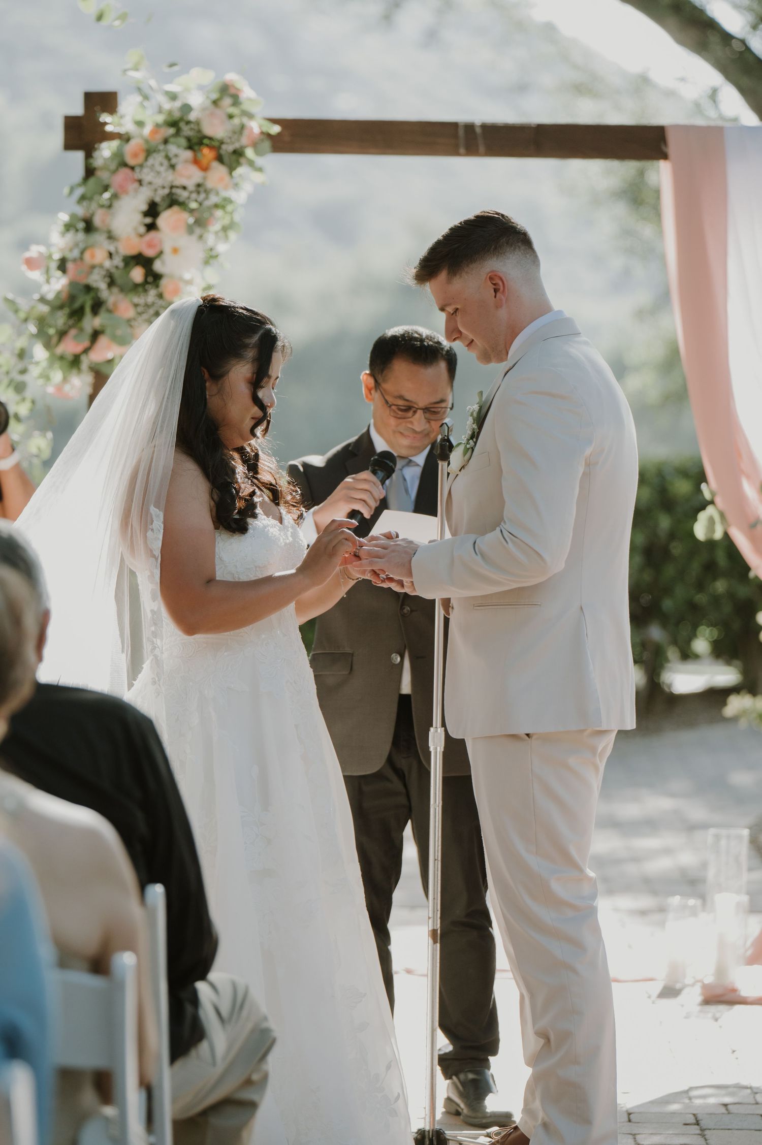 Bride places wedding ring on groom's finger during outdoor ceremony at floral arch with officiant.