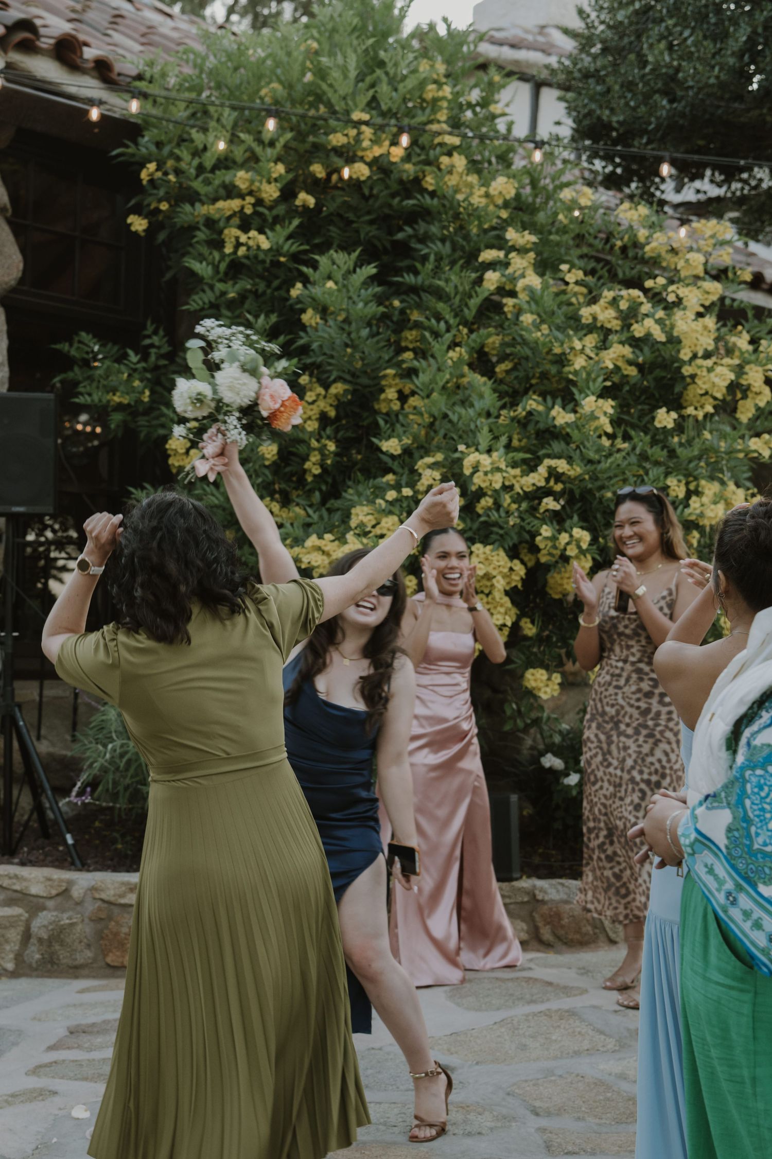 Woman in green dress catches bridal bouquet as guests celebrate at an outdoor wedding reception.