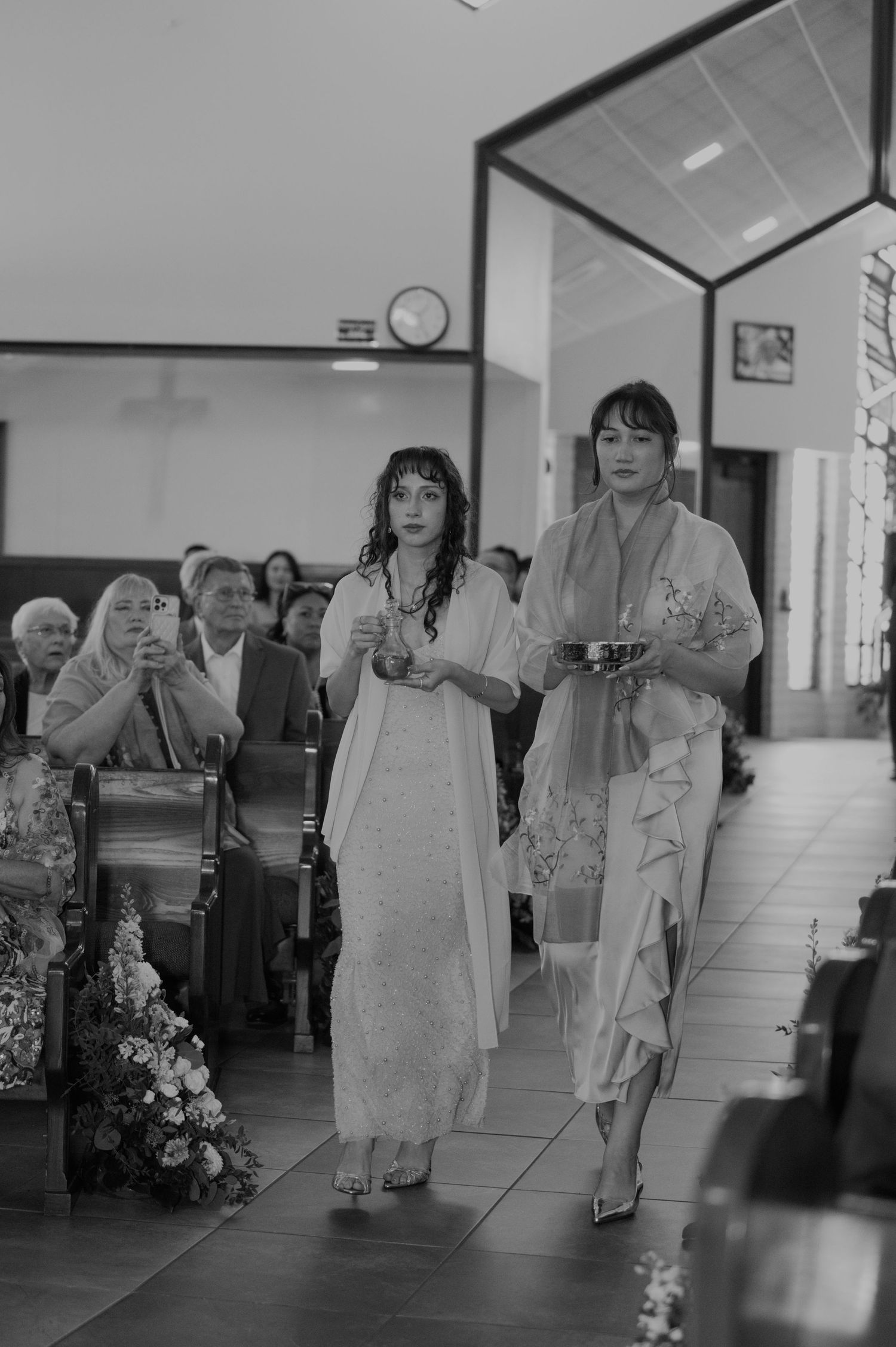 Two elegantly dressed women walk down a church aisle during a wedding ceremony in black and white.