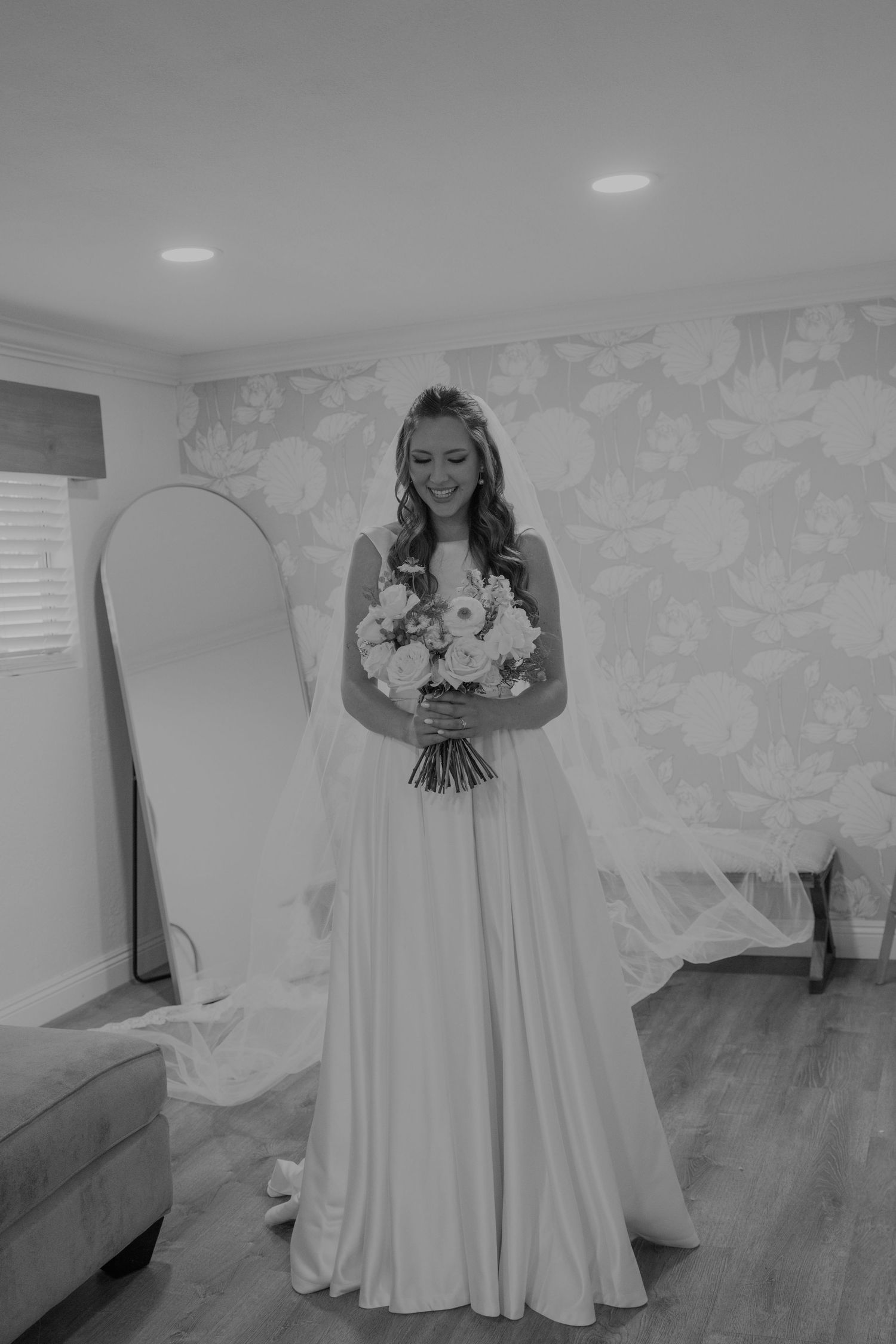 Smiling bride in elegant white gown holds bouquet in floral-wallpapered bridal suite, black and white photo.