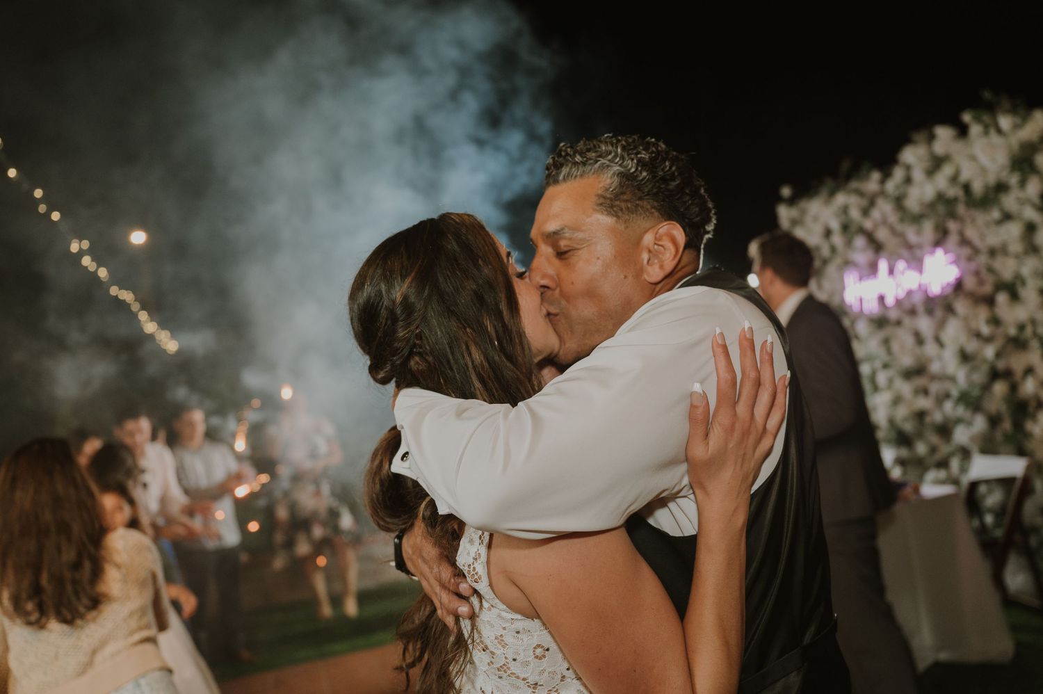 Couple sharing a romantic kiss during wedding reception with sparklers and floral backdrop behind them.
