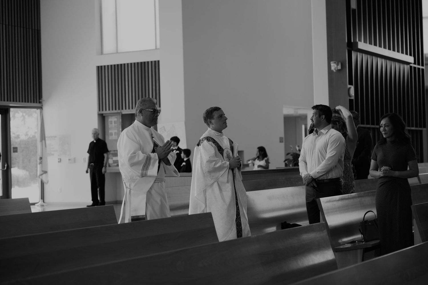 Priests and guests gather in a modern church before a wedding ceremony in black and white photo.