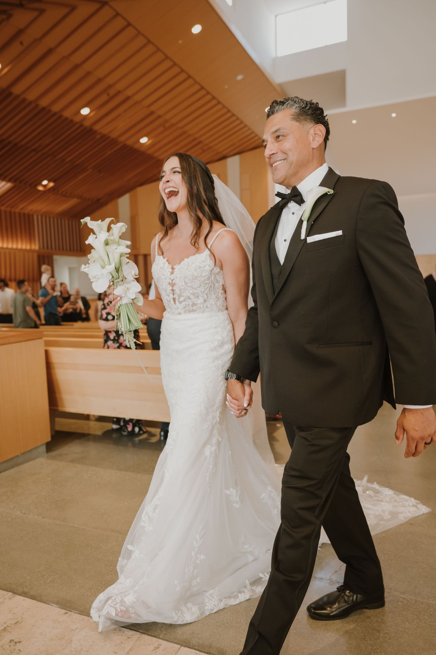 Joyful newlyweds walk down church aisle, bride in lace gown holding white bouquet, groom in black tuxedo.