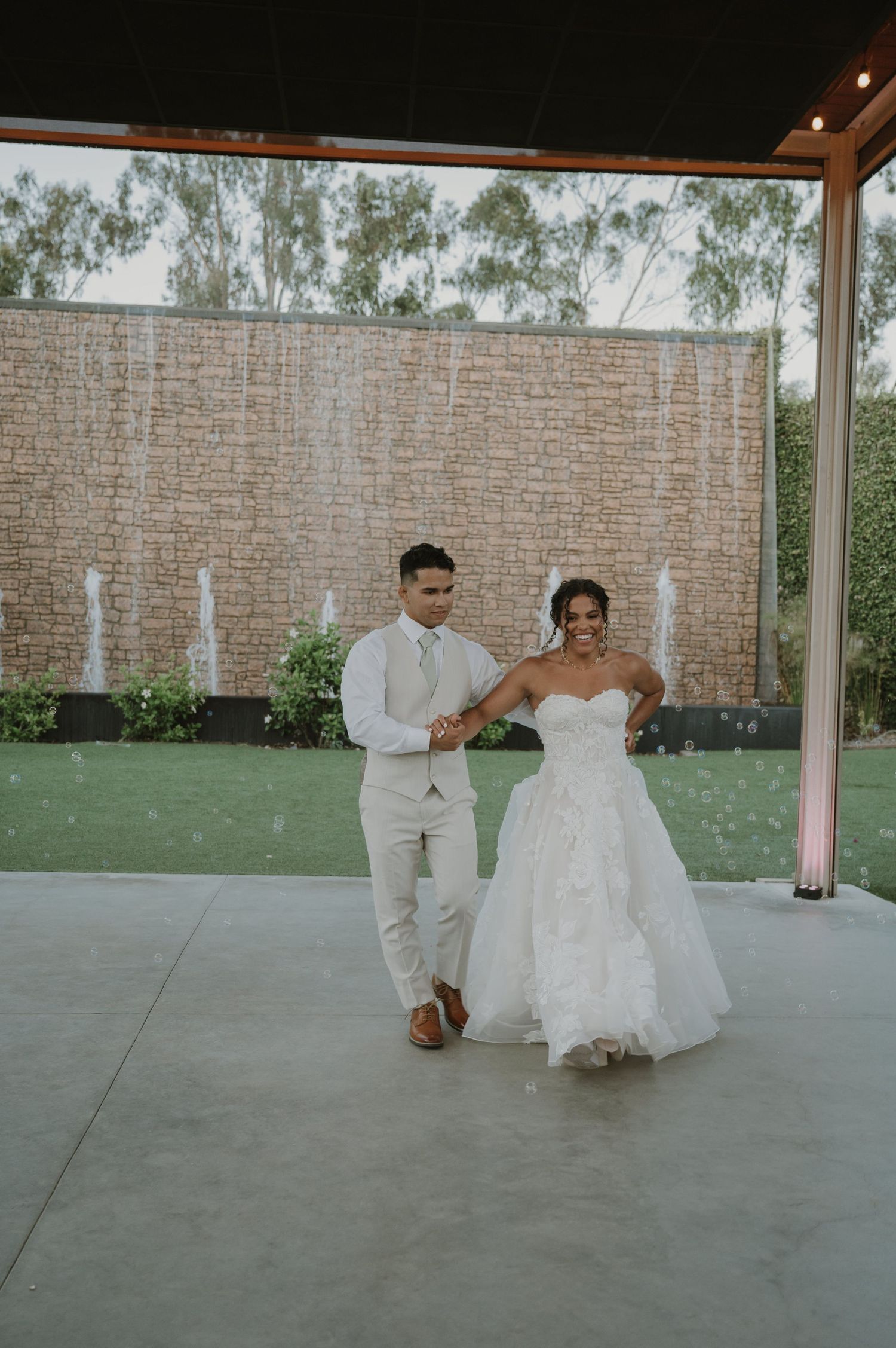 Bride in strapless white gown and groom in beige suit share first dance outdoors at elegant wedding venue with water wall.