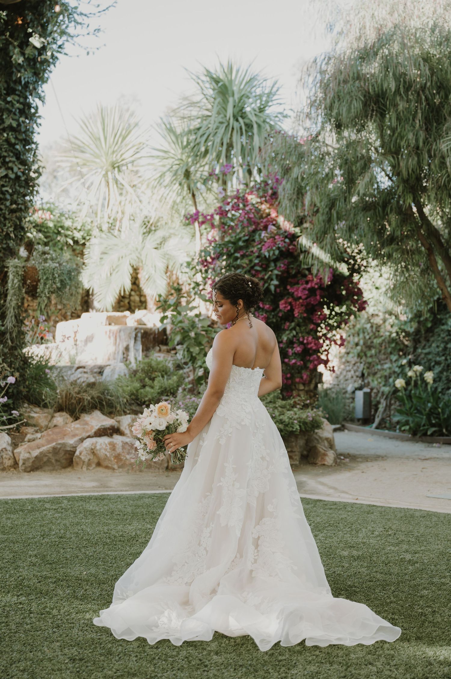 Bride in strapless lace wedding gown holding bouquet in lush garden with tropical plants and colorful flowers.