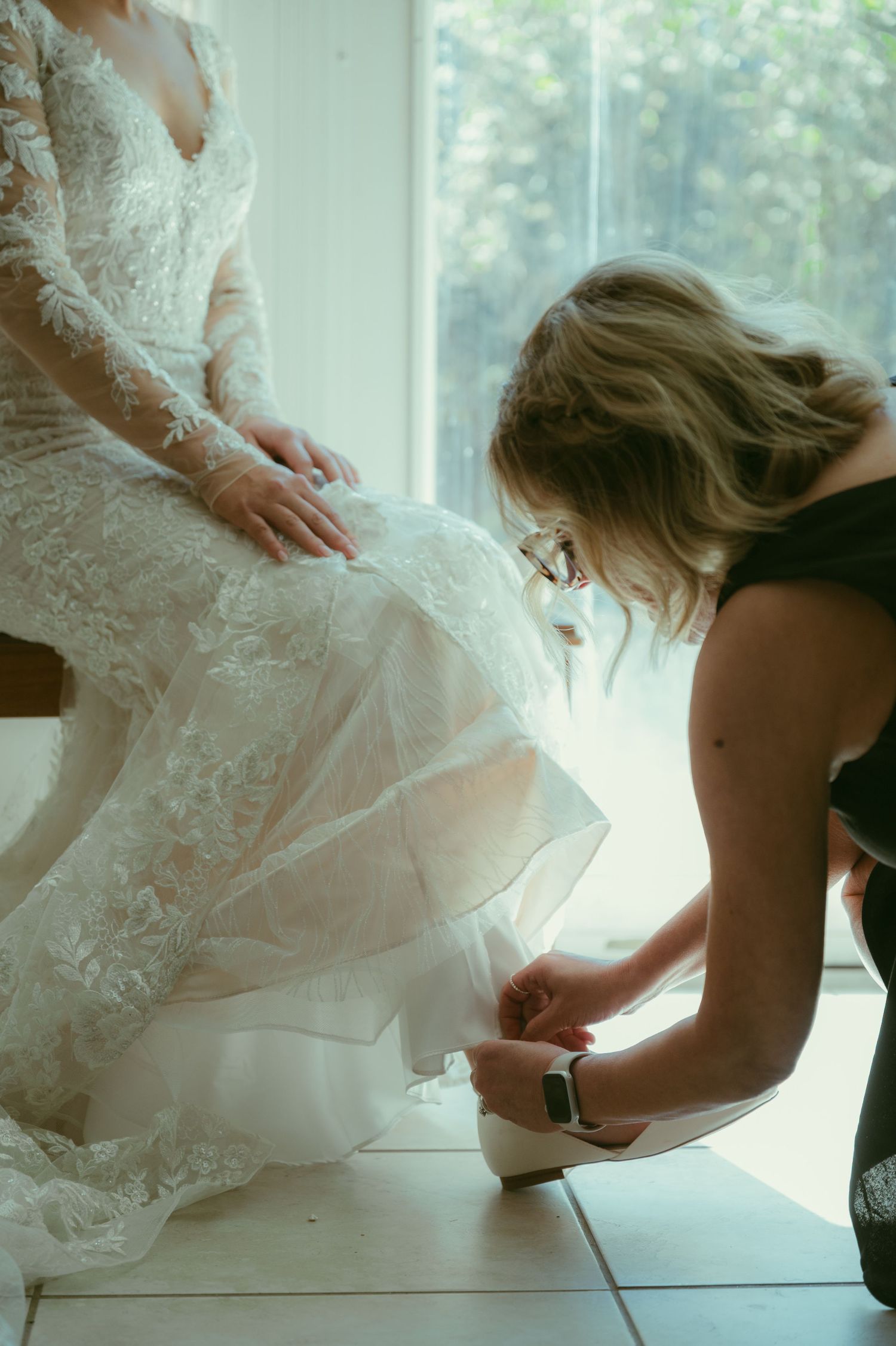 Bridesmaid kneeling to help the bride put on her white wedding heels before the ceremony.