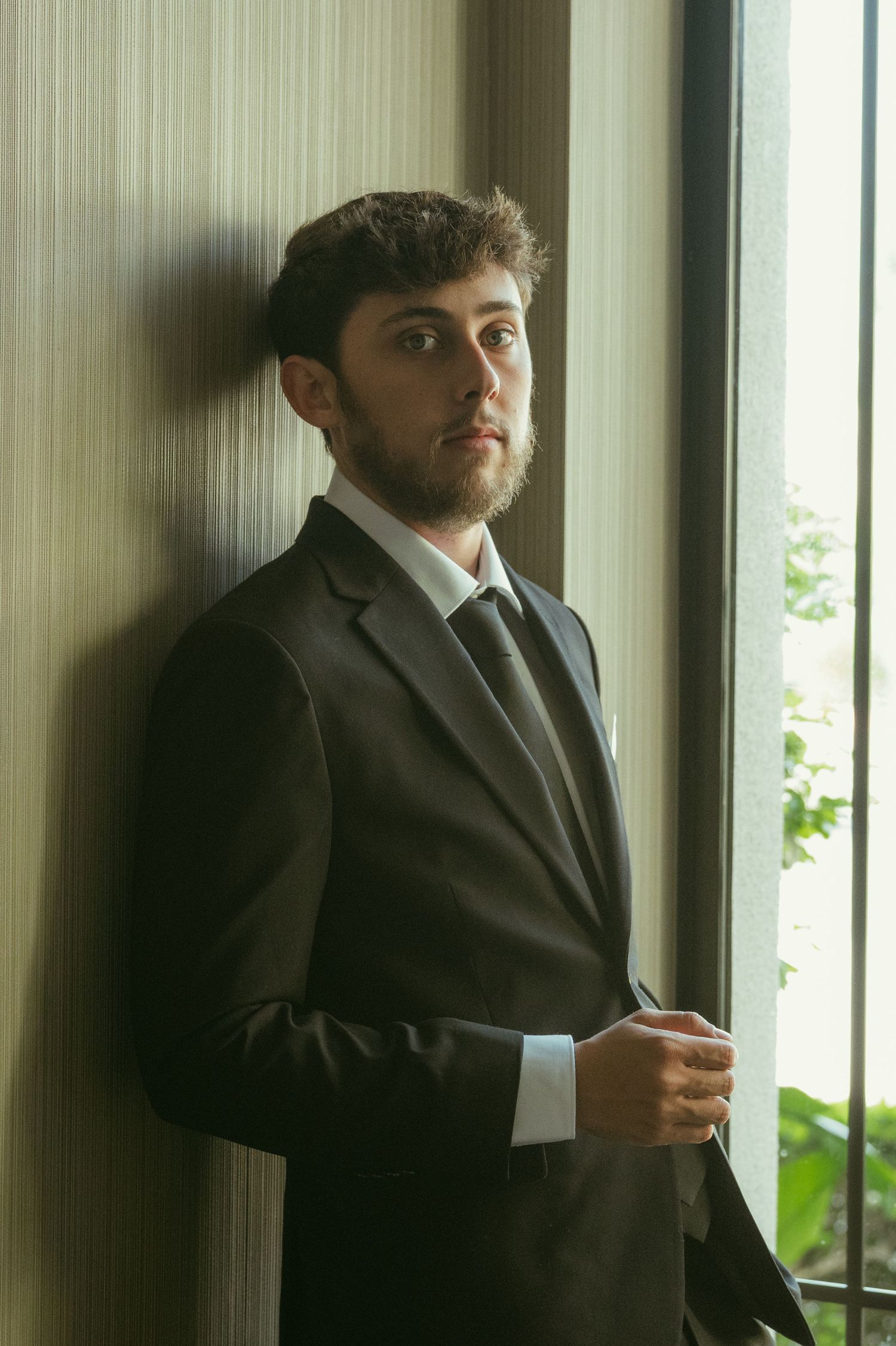 Groom in dark suit and tie leaning against wall by window with natural light.