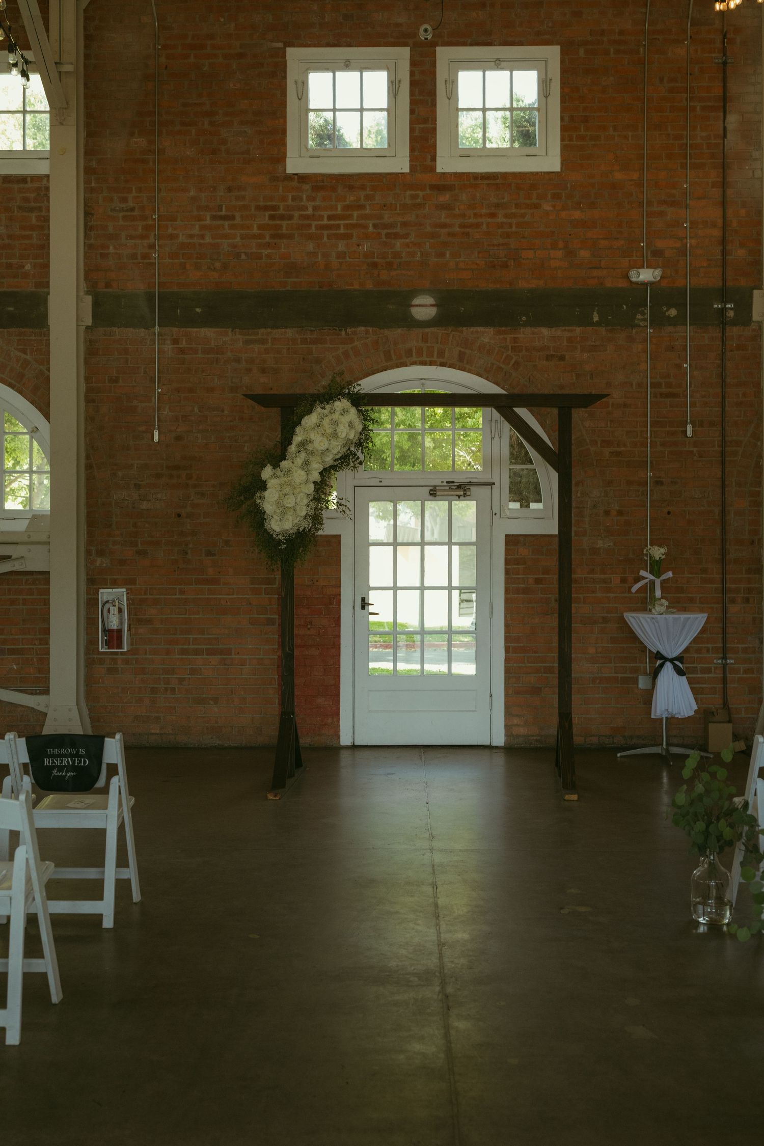 Rustic brick wedding venue interior with floral arch, white chairs, and elegant decor for a ceremony setup.