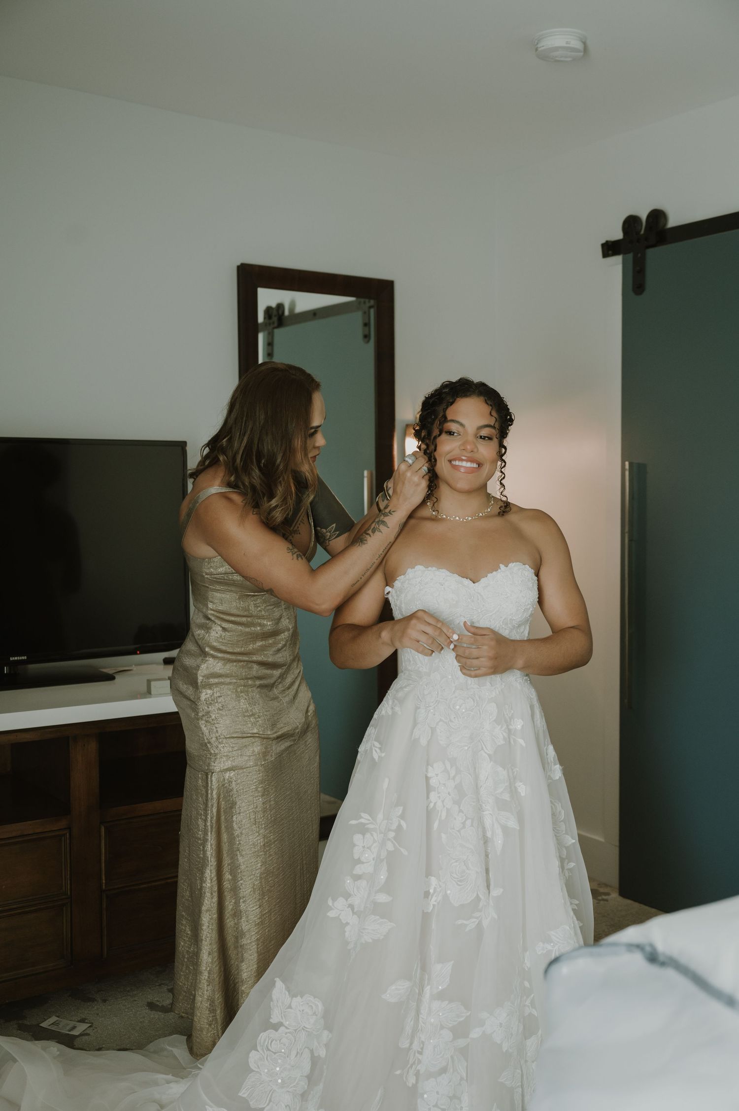 Mother helping bride into her floral lace strapless wedding dress in a hotel room before the ceremony.