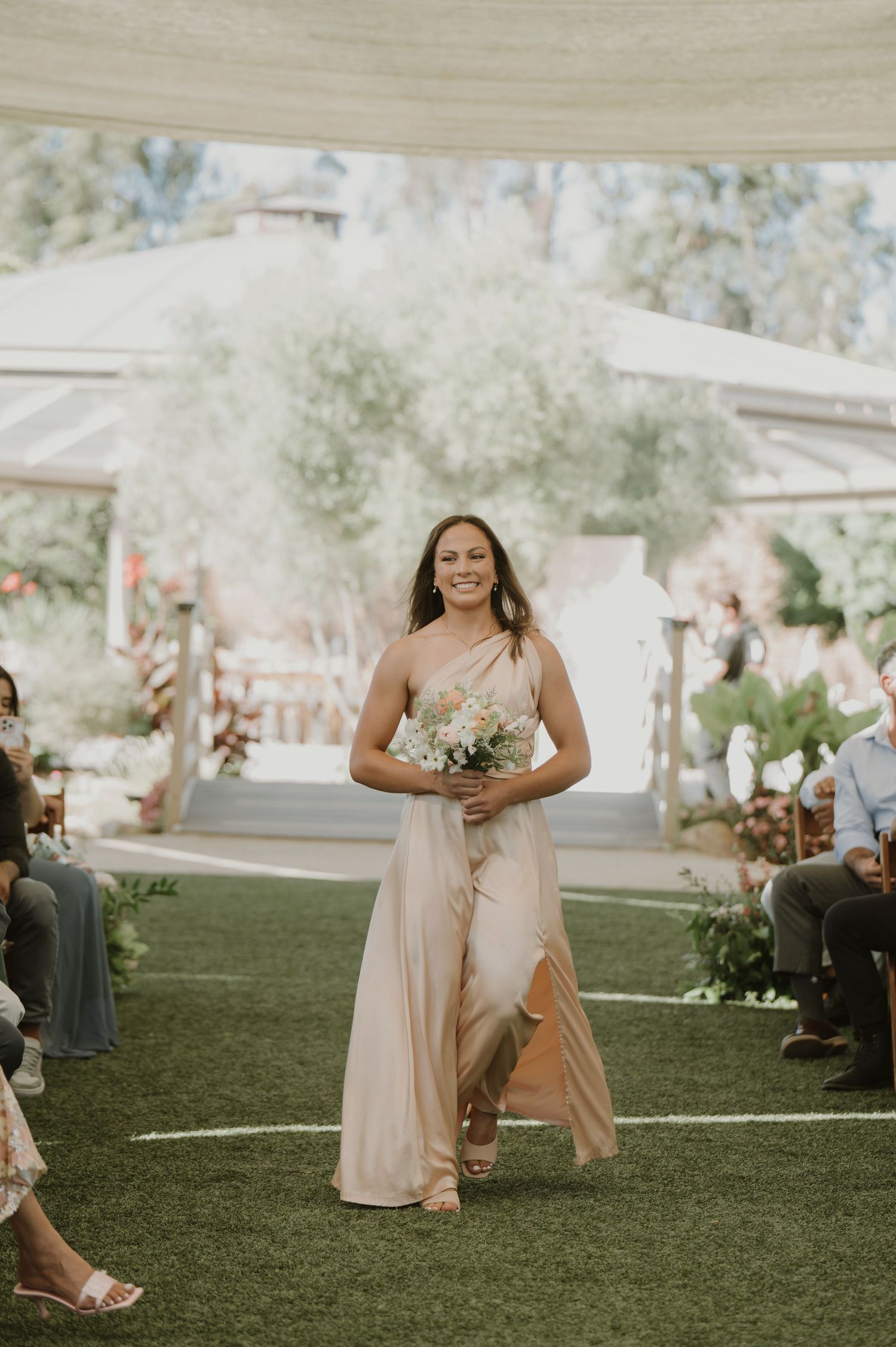 Bridesmaid in a blush one-shoulder gown walks down the aisle holding a pink floral bouquet at an outdoor wedding.