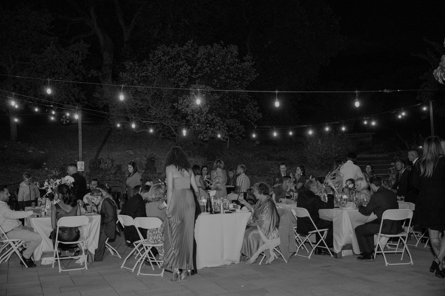 Black and white outdoor wedding reception at night with string lights, guests seated at round tables on a patio.