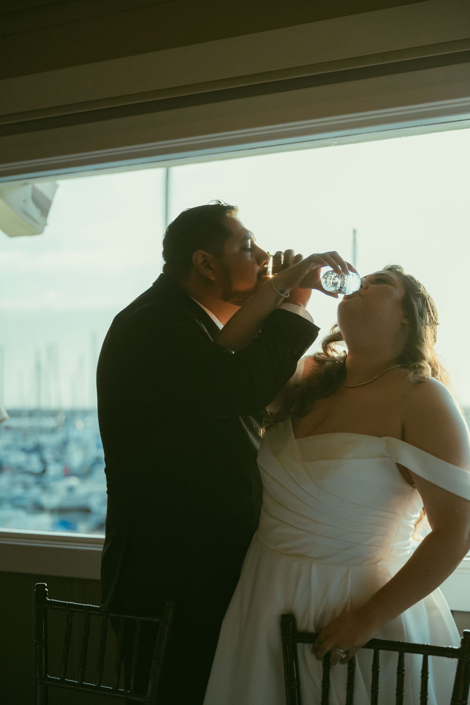 Bride and groom drinking from glasses at wedding reception near large window with marina view.