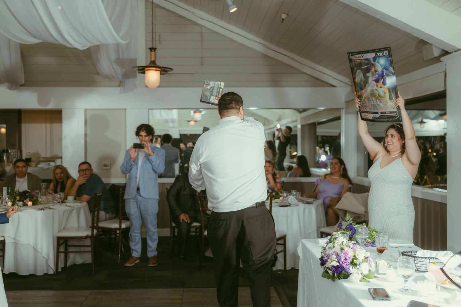 Bride and groom holding framed photos at wedding reception while guests celebrate at decorated tables.
