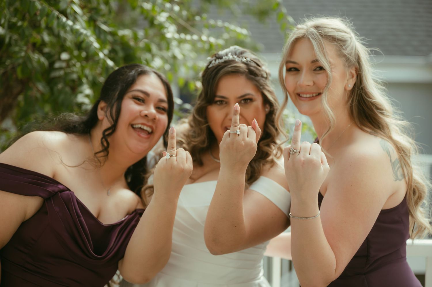 A bride in white and two bridesmaids in purple dresses playfully show off their rings outdoors.