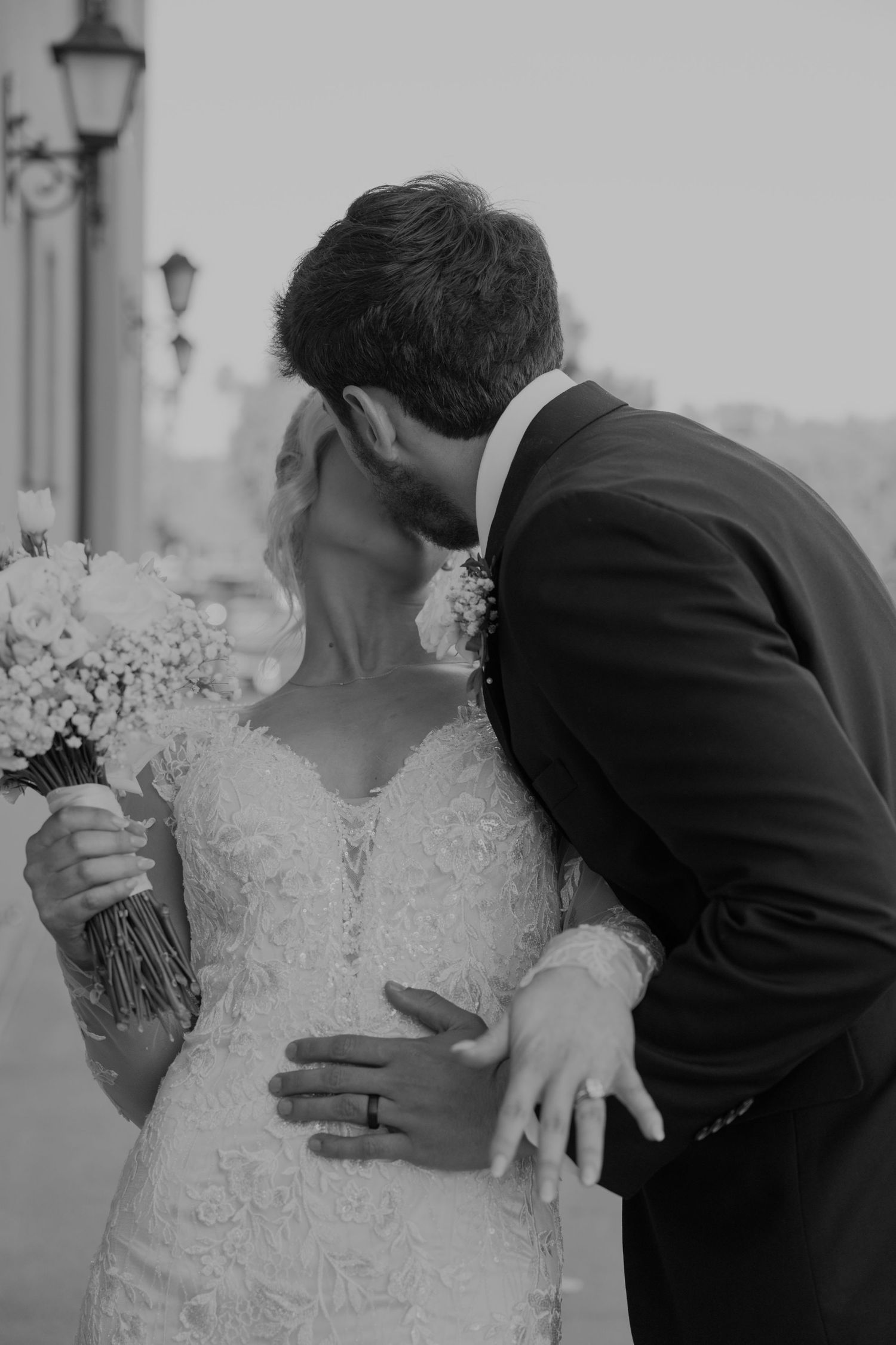 Black and white photo of a bride and groom sharing a kiss, bride holding a floral bouquet in a lace wedding dress.
