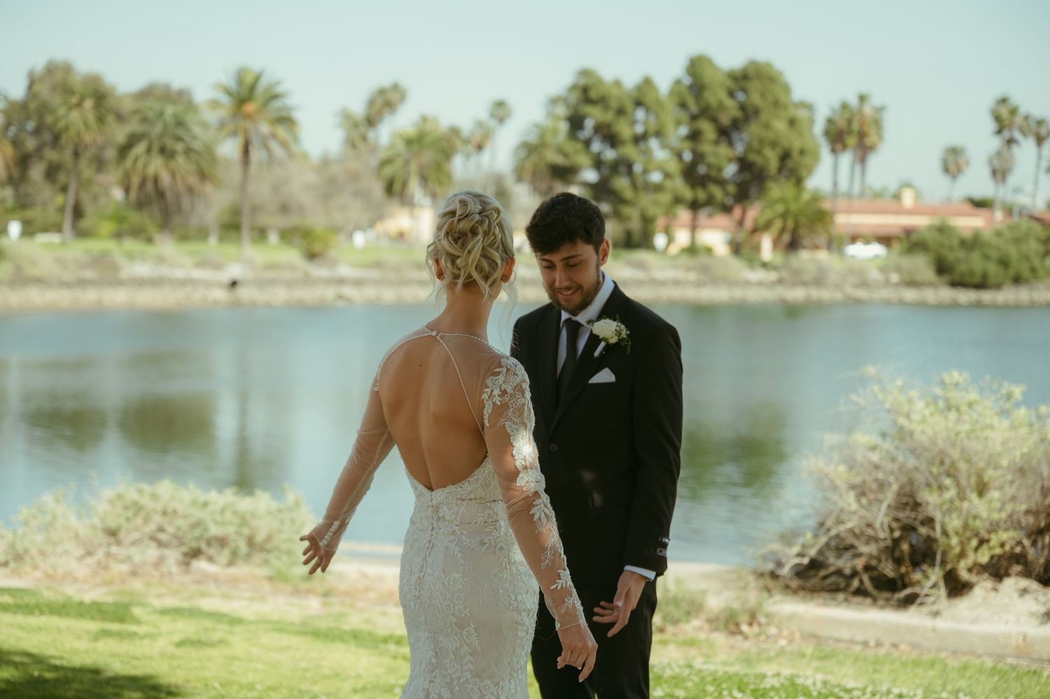 Bride and groom share romantic first look moment holding hands by serene lake with palm trees backdrop.