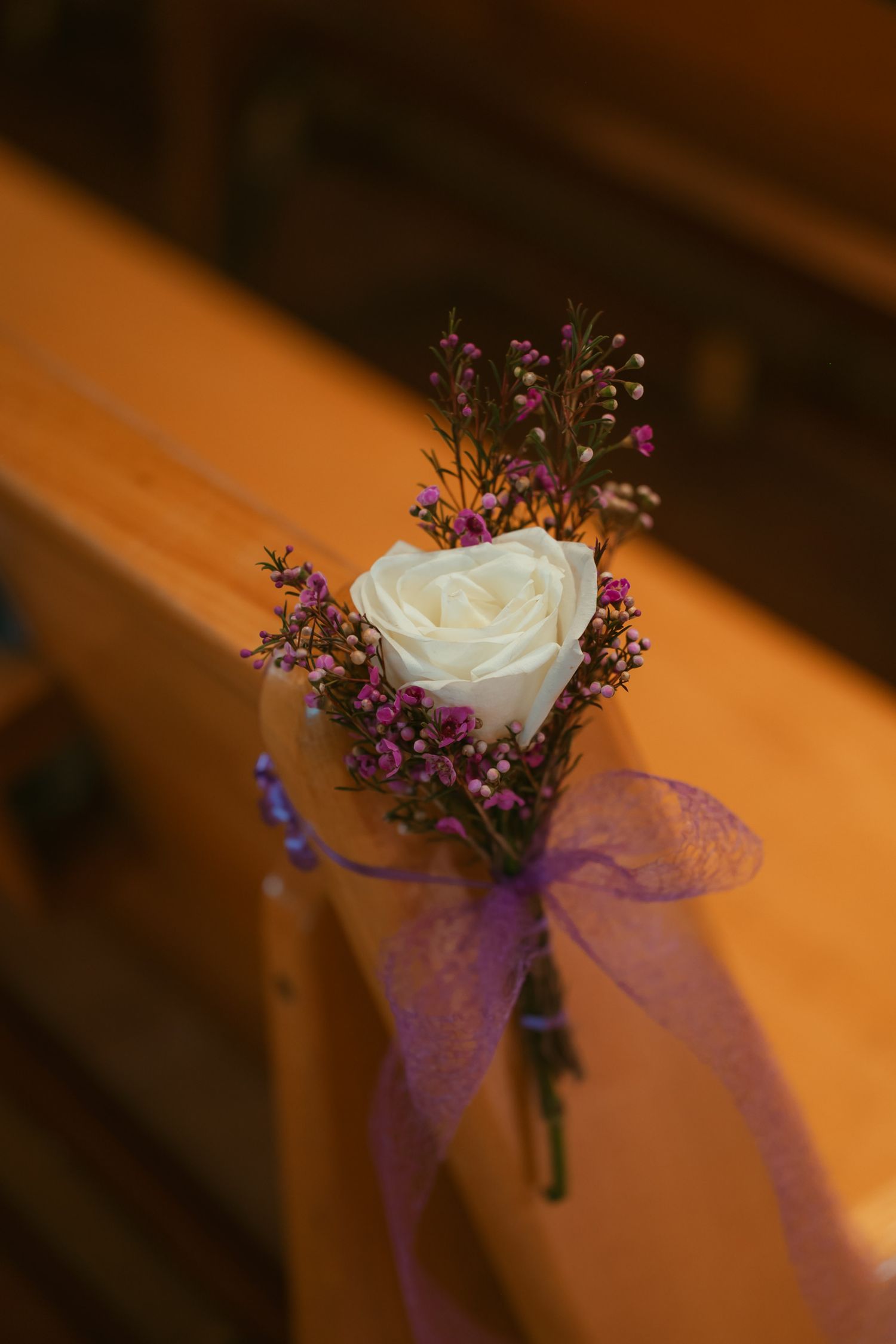 White rose boutonniere with purple wildflowers and ribbon tied to wooden church pew.