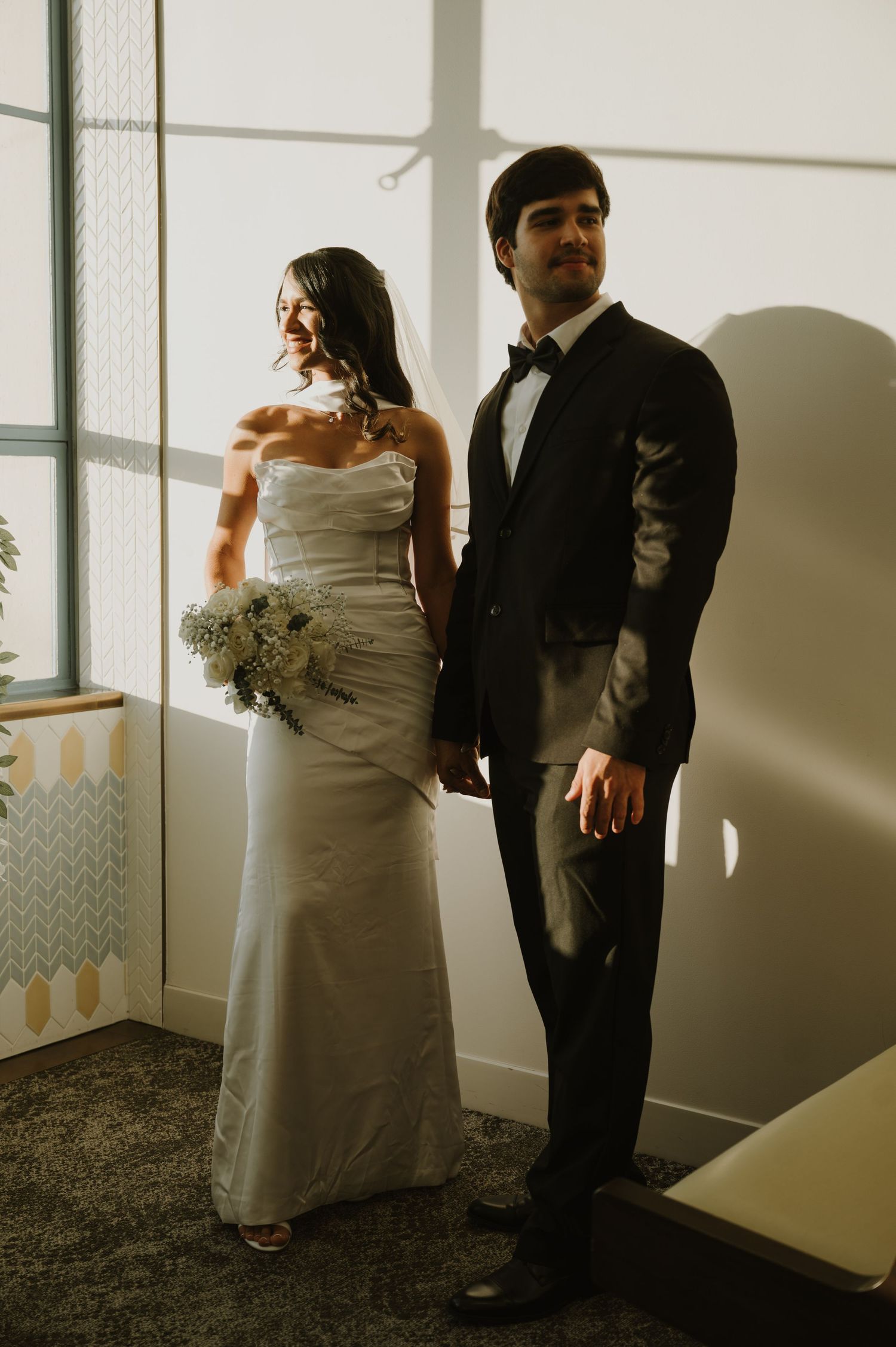 Wedding couple poses near sunlit windows in a bright indoor space.