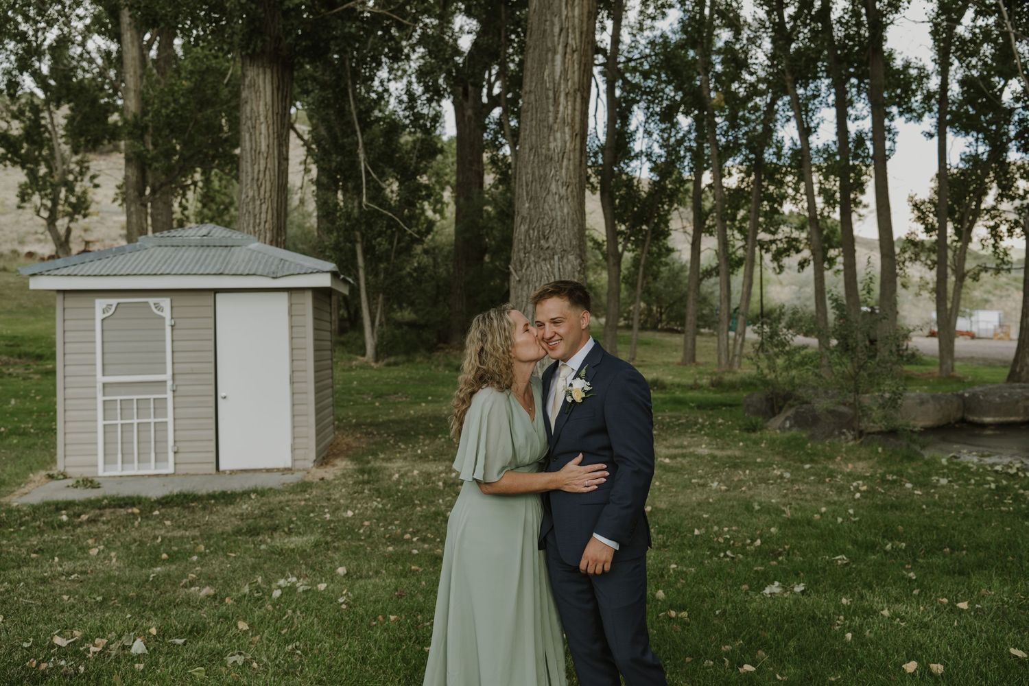 A couple poses together on a grassy lawn with trees and a small building in the background.