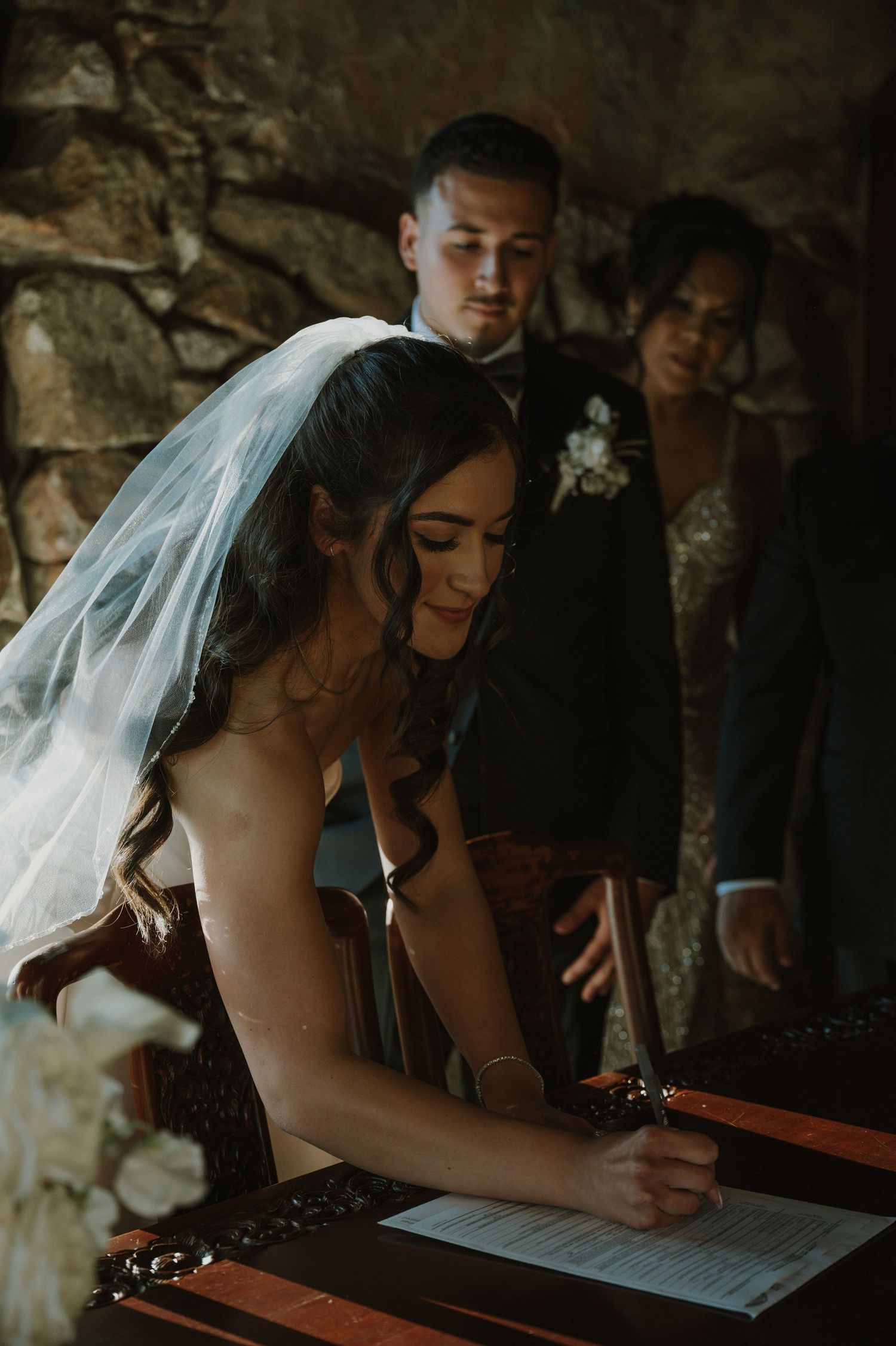 A bride in white dress signing wedding documents in a rustic stone interior.