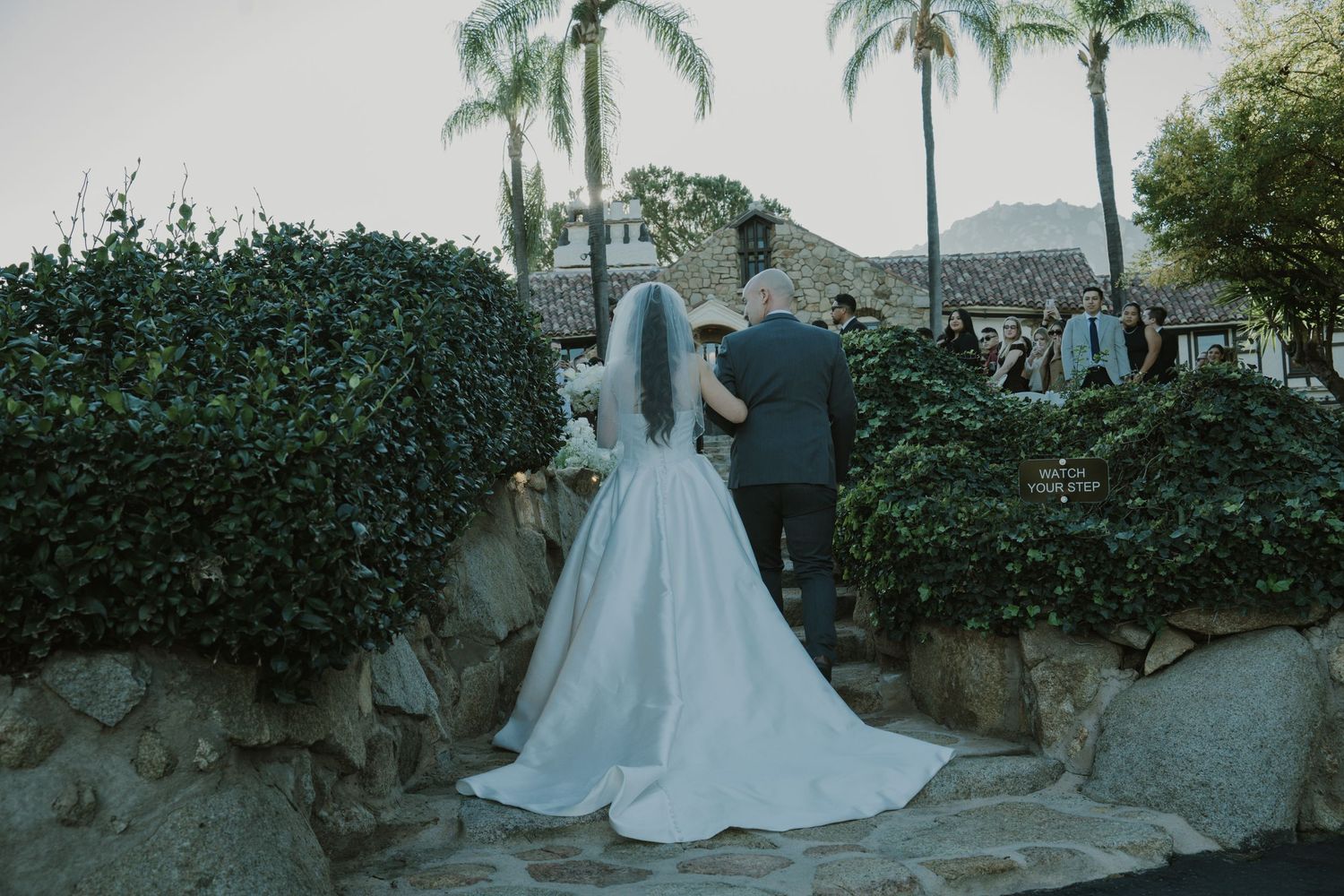 Bride in flowing white gown walking through garden setting.
