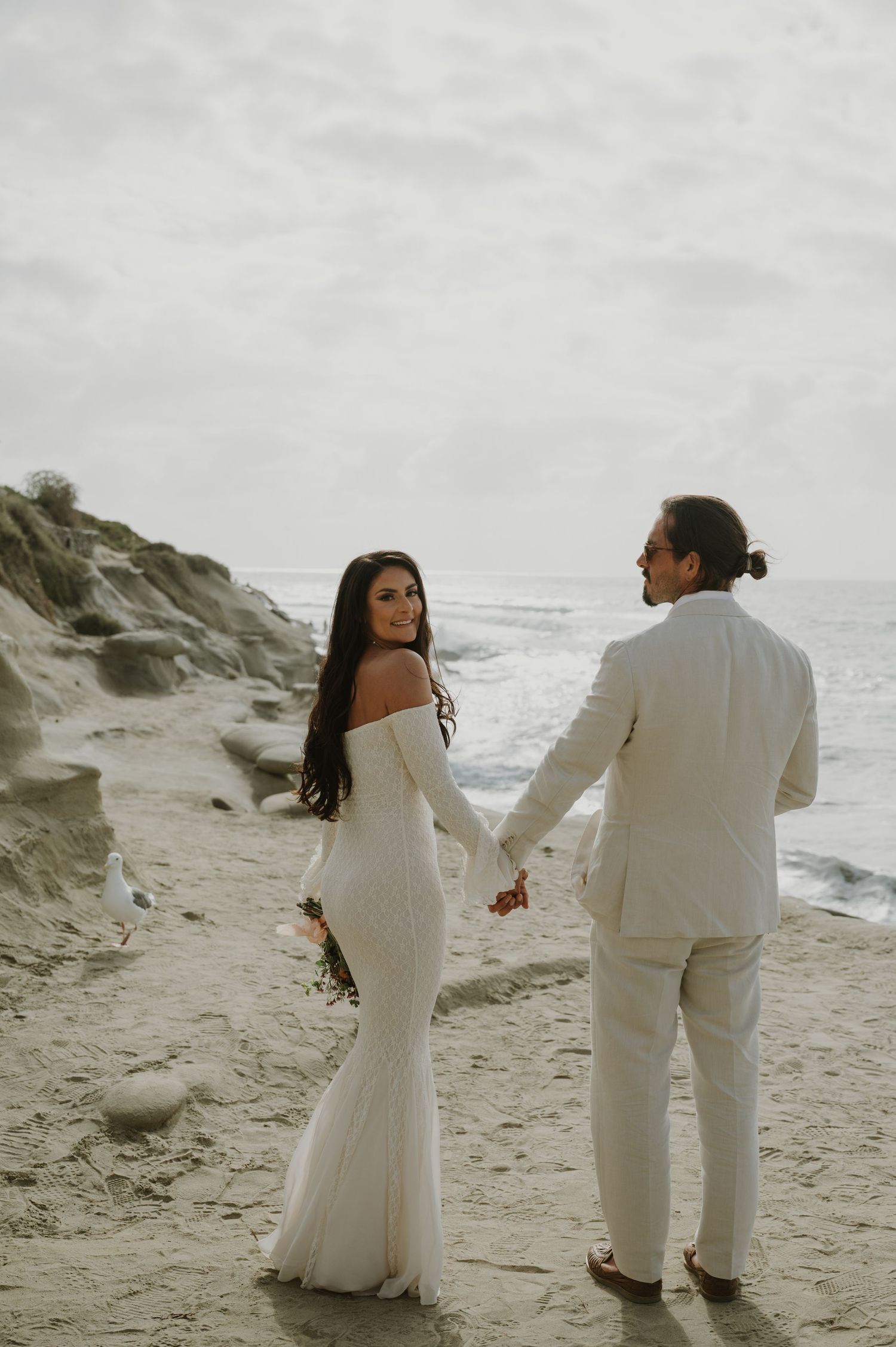 A couple in matching white outfits walks playfully along a sandy beach.