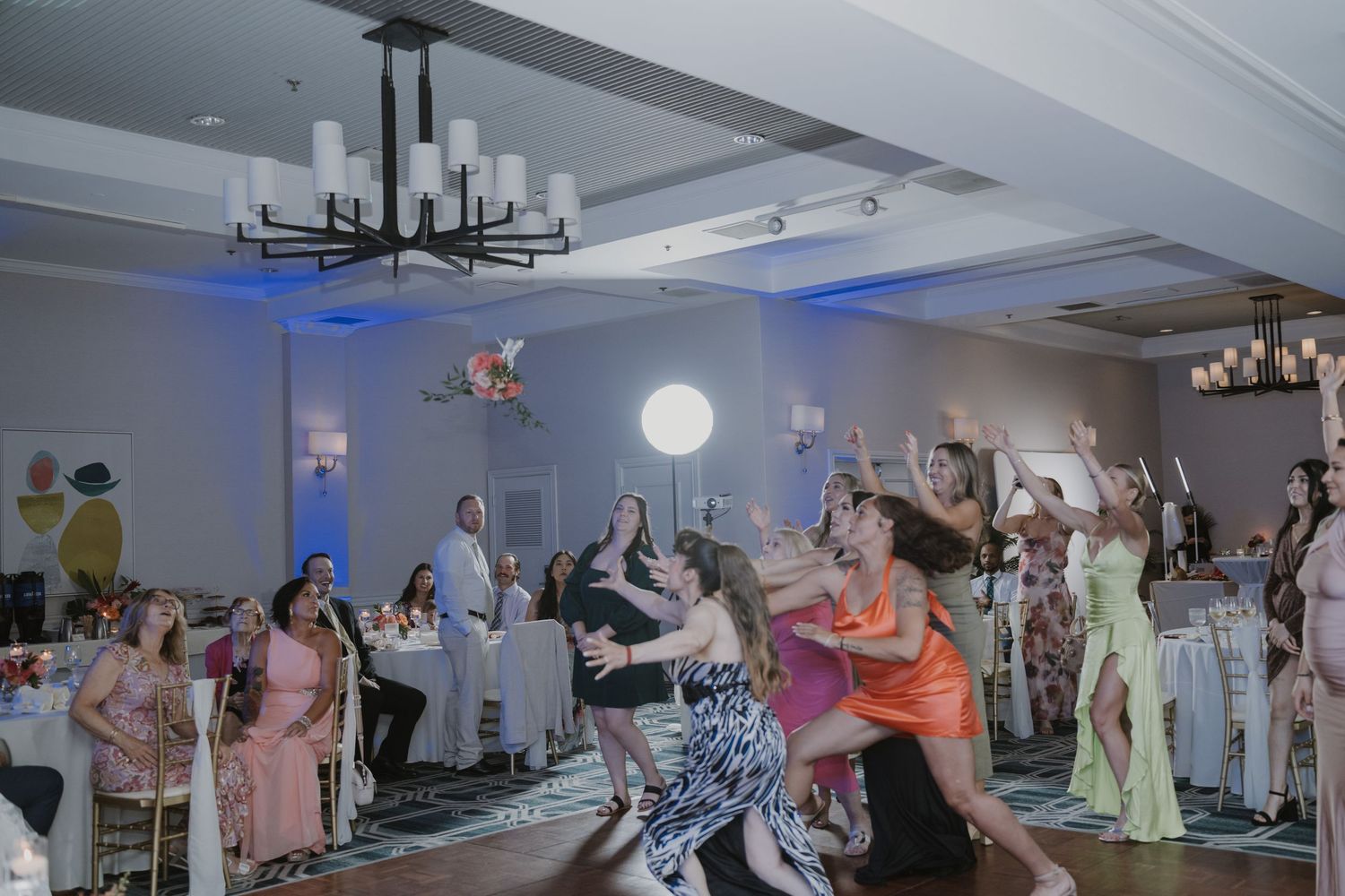 Wedding reception venue with modern chandeliers and guests dancing on parquet flooring.