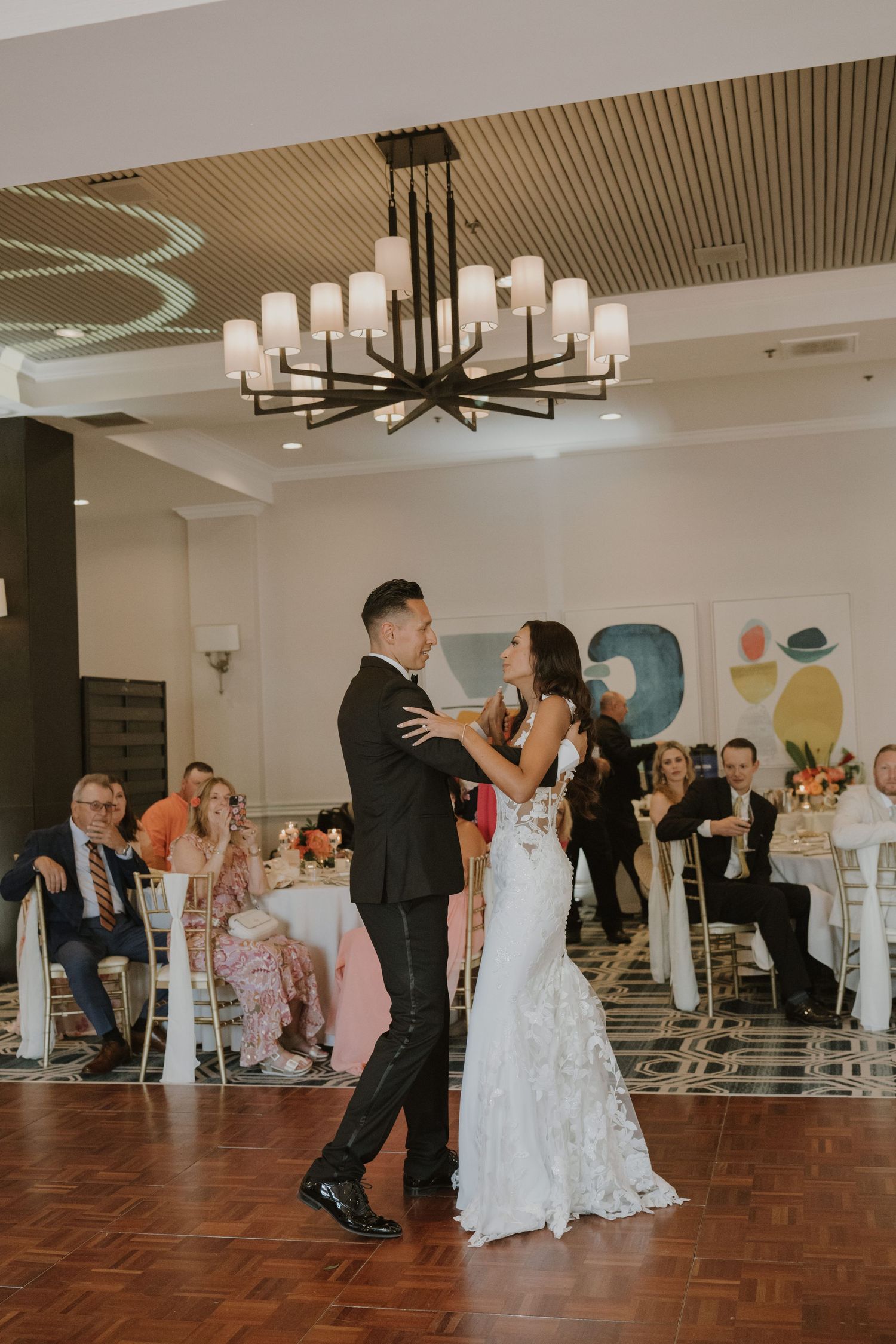 A couple shares their first dance under an elegant chandelier surrounded by wedding guests.