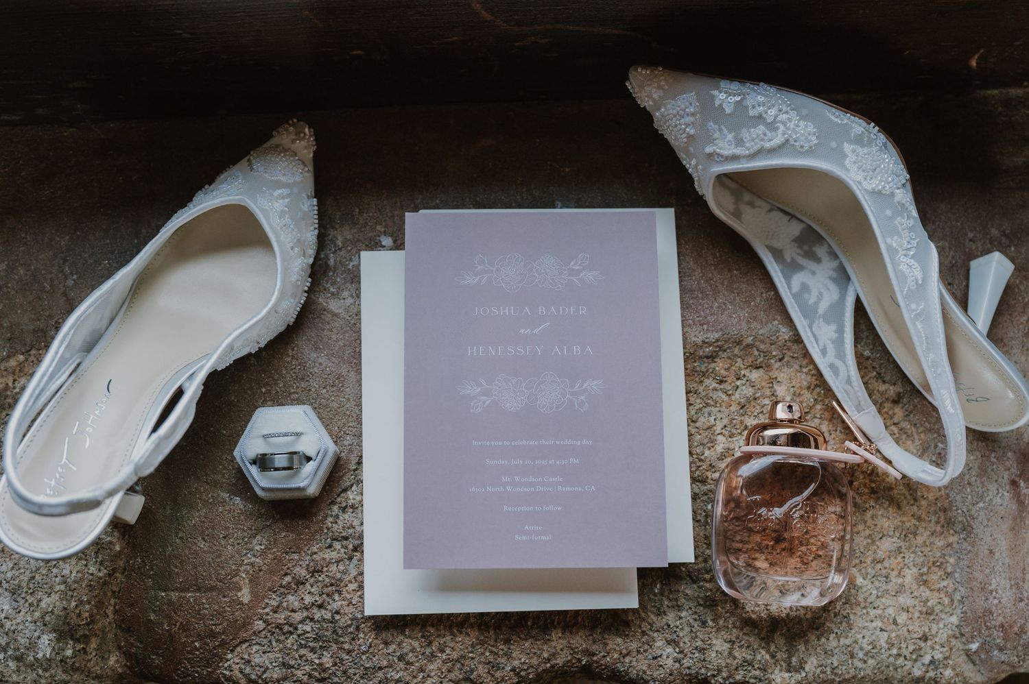 Elegant wedding flat lay featuring lace heels, floral invitation suite, ring box, and perfume bottle on stone surface.